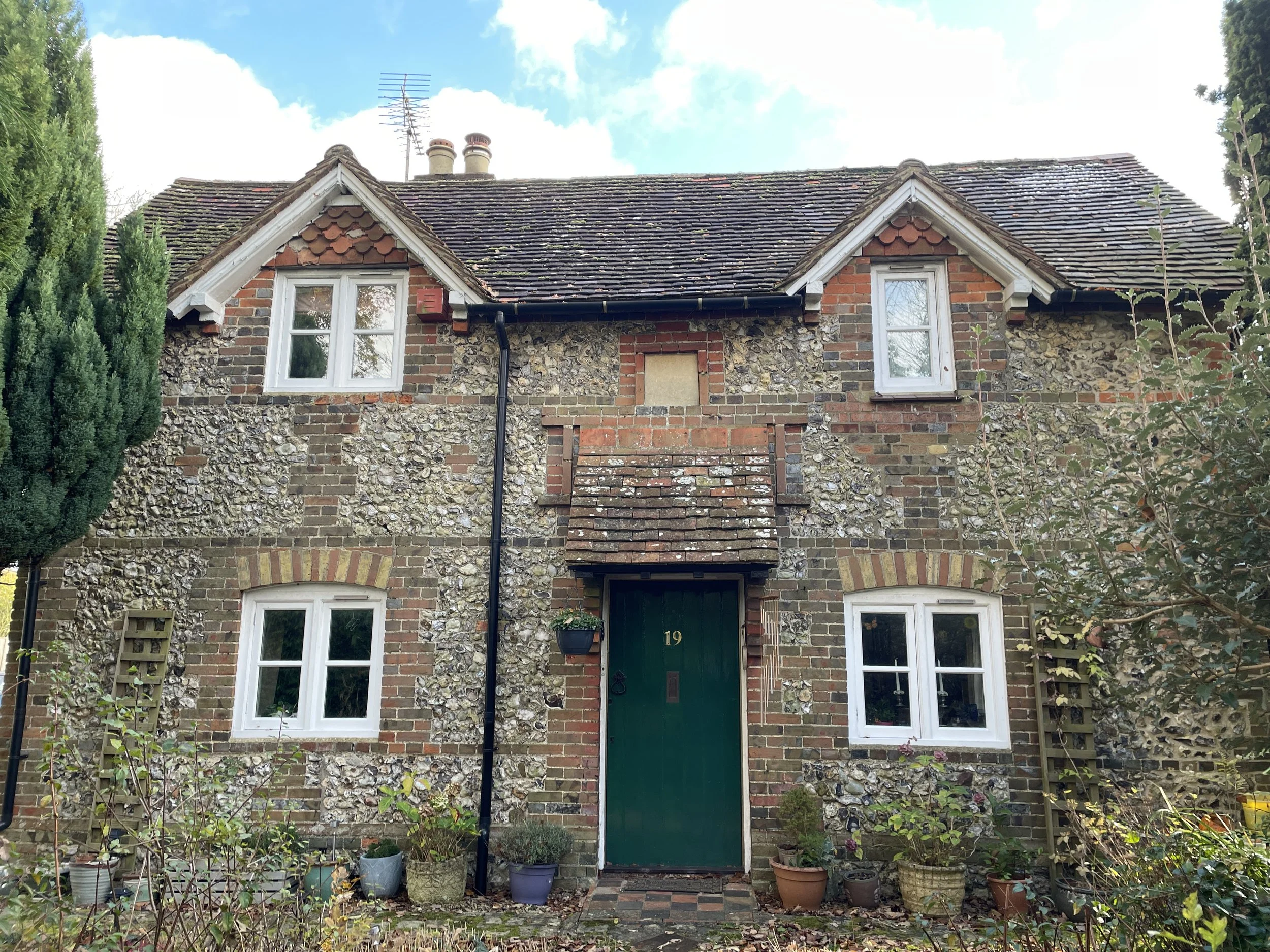 Front view of a two-story house with brick and flint exterior, white-framed windows, green door with the number 19, potted plants near entrance, and a garden with bushes and trees.