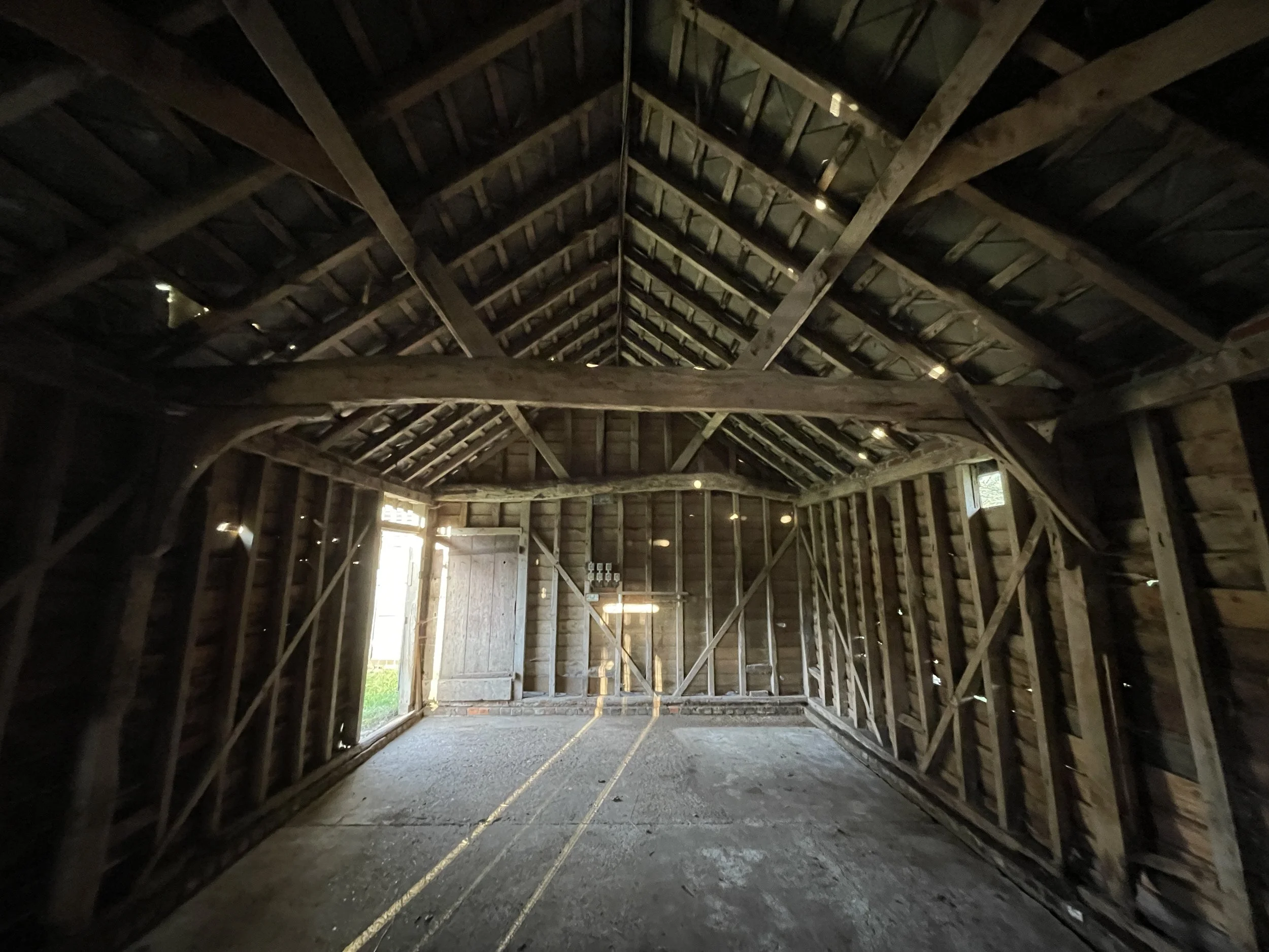 Interior of a wooden barn with exposed beams and siding, unfinished floor, and natural light coming from open window and door.