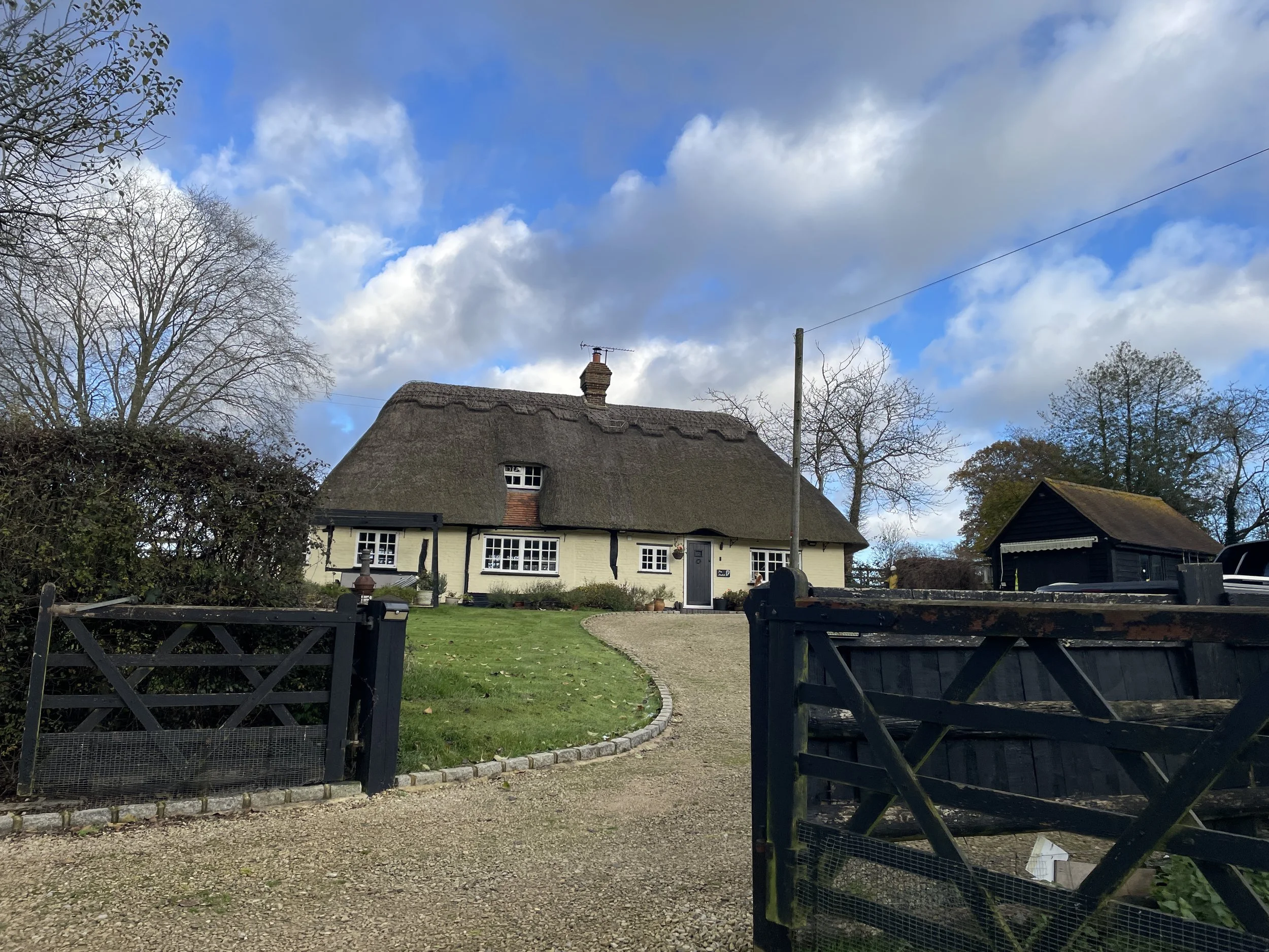 A cottage with a thatched roof, white walls, black trim, and a small front garden with a curved path leading to the door. There are leafless trees and a partly cloudy sky in the background.
