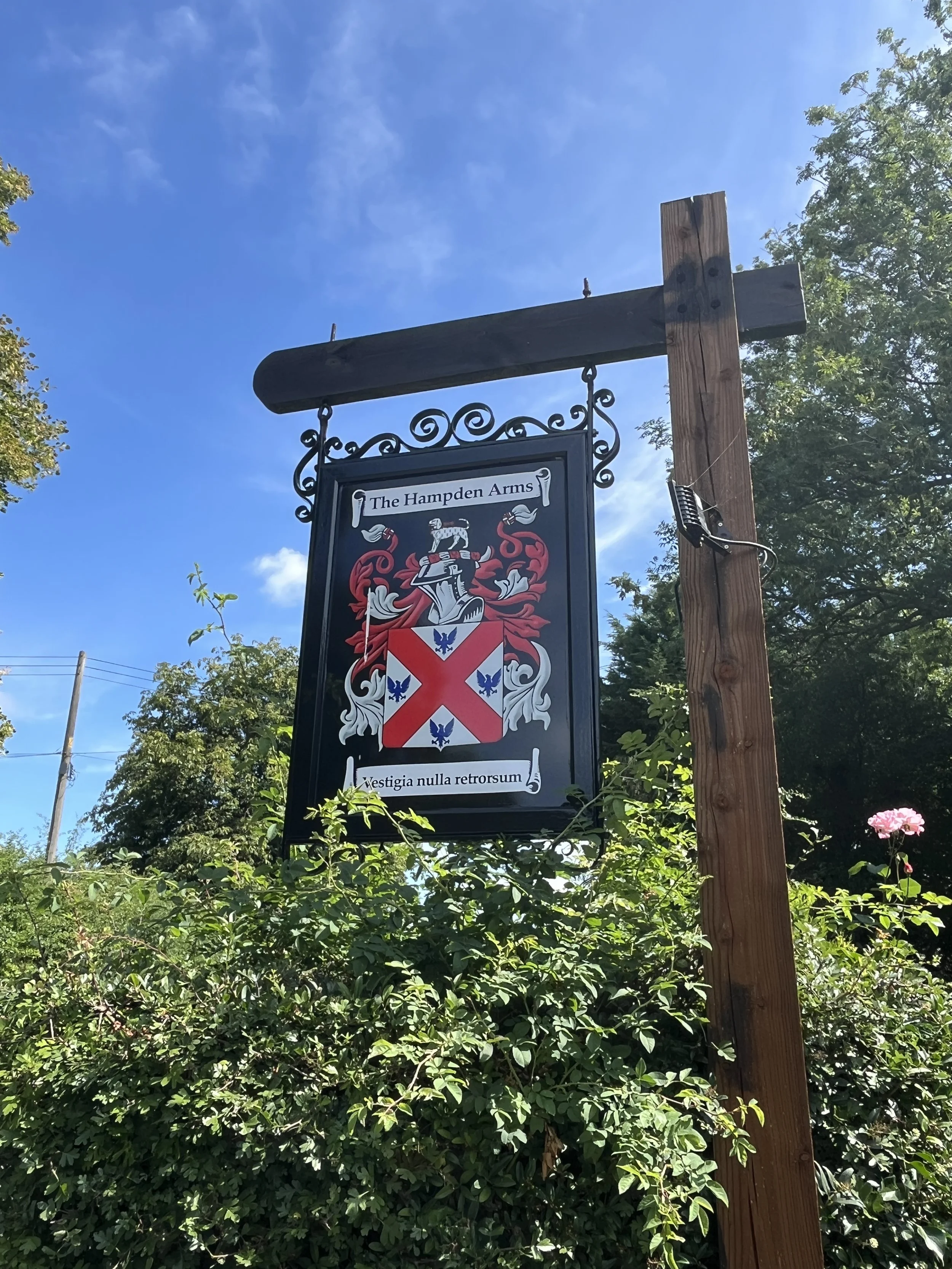 Sign for The Hampden Arms pub with a coat of arms and Latin phrase, mounted on a wooden post against a blue sky and surrounded by green foliage.