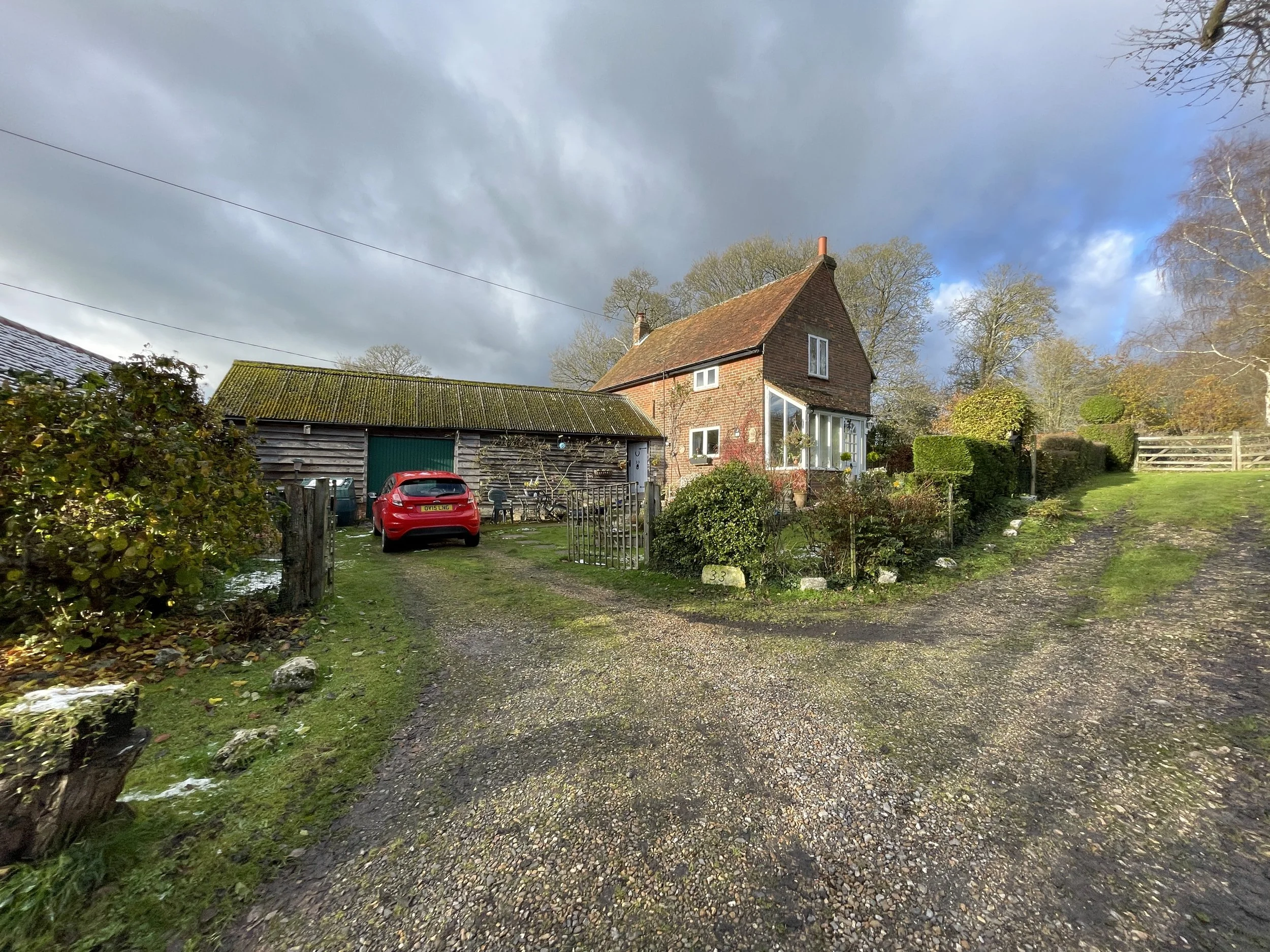 A country house with red brick walls and a steep roof, surrounded by a garden with bushes and trees. There's a driveway with a red car parked and a wooden garage in the background. The sky is partly cloudy with patches of blue.