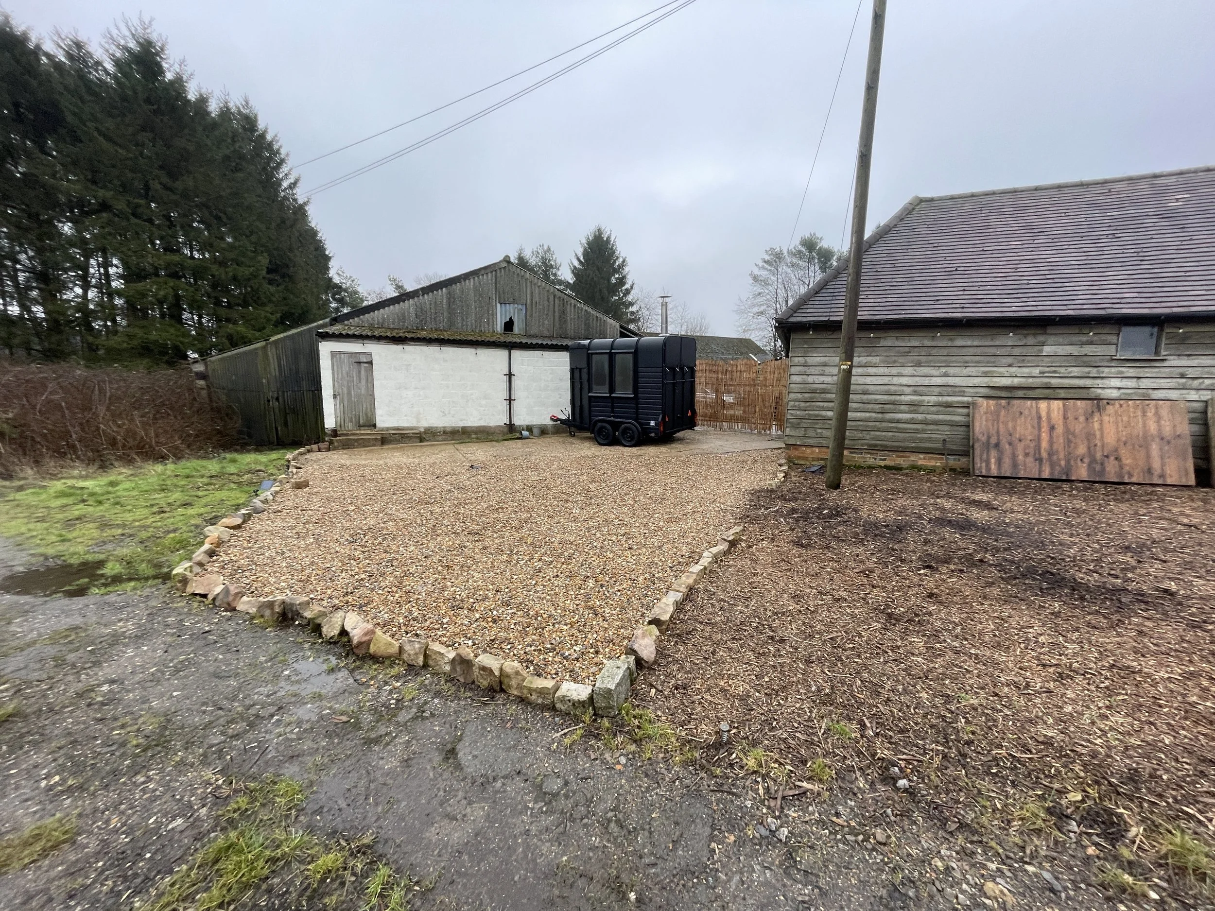 A gravel driveway bordered with bricks leading to a white shed, with a small black trailer parked nearby, adjacent to a weathered wooden building, overcast weather, and some trees in the background.