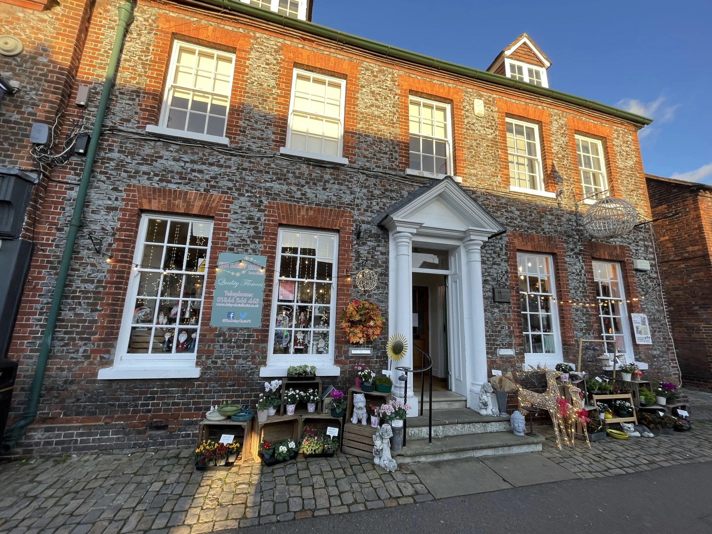 A brick storefront with three floors, decorated with holiday lights and ornaments, featuring a sign for a flower shop with potted plants and seasonal decorations outside.