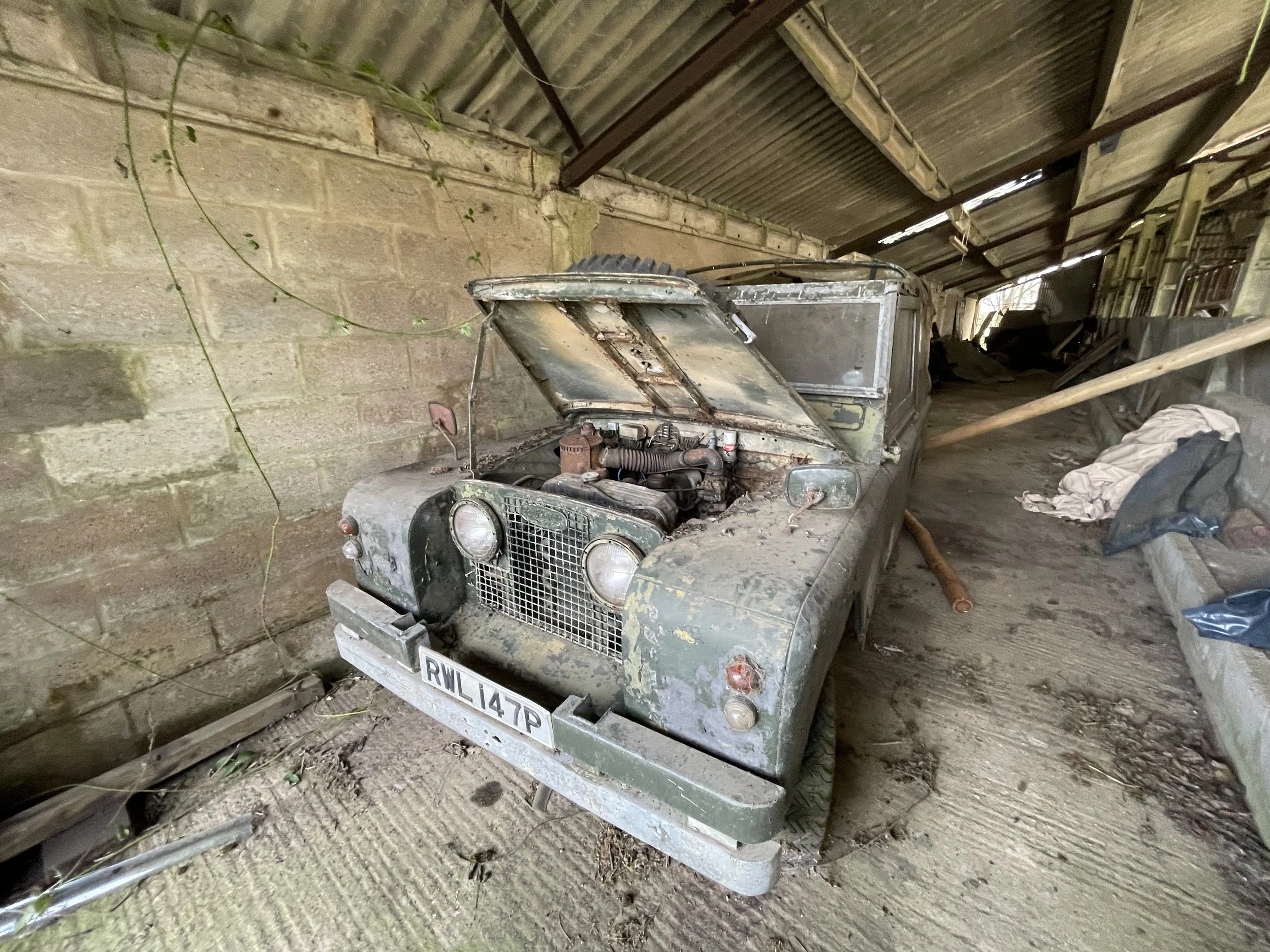 An old, dusty vintage vehicle with an open hood in a barn-like storage area, surrounded by clutter and debris.