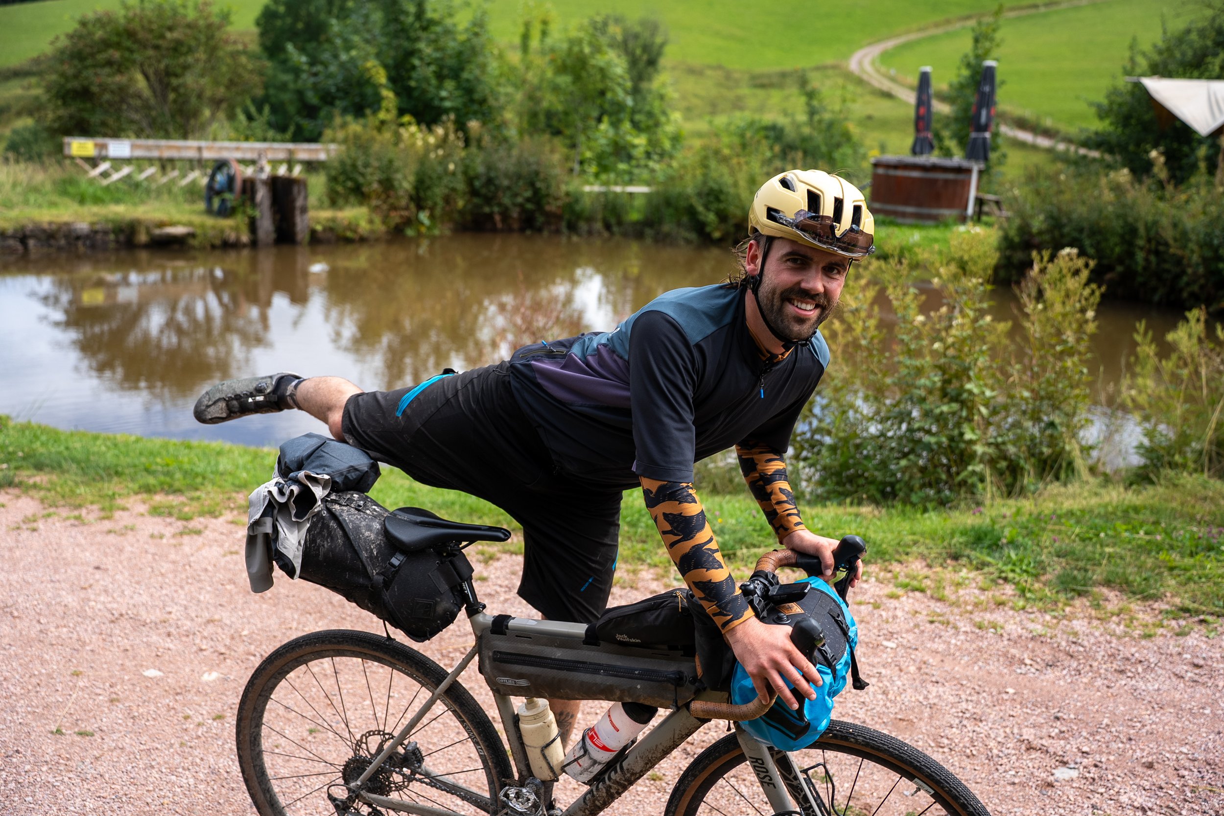 Ein Mann in Radfahrmontur mit Helm und Arm- und Beinschützern, der auf einem Fahrrad im Freien neben einem kleinen See liegt und lächelt.