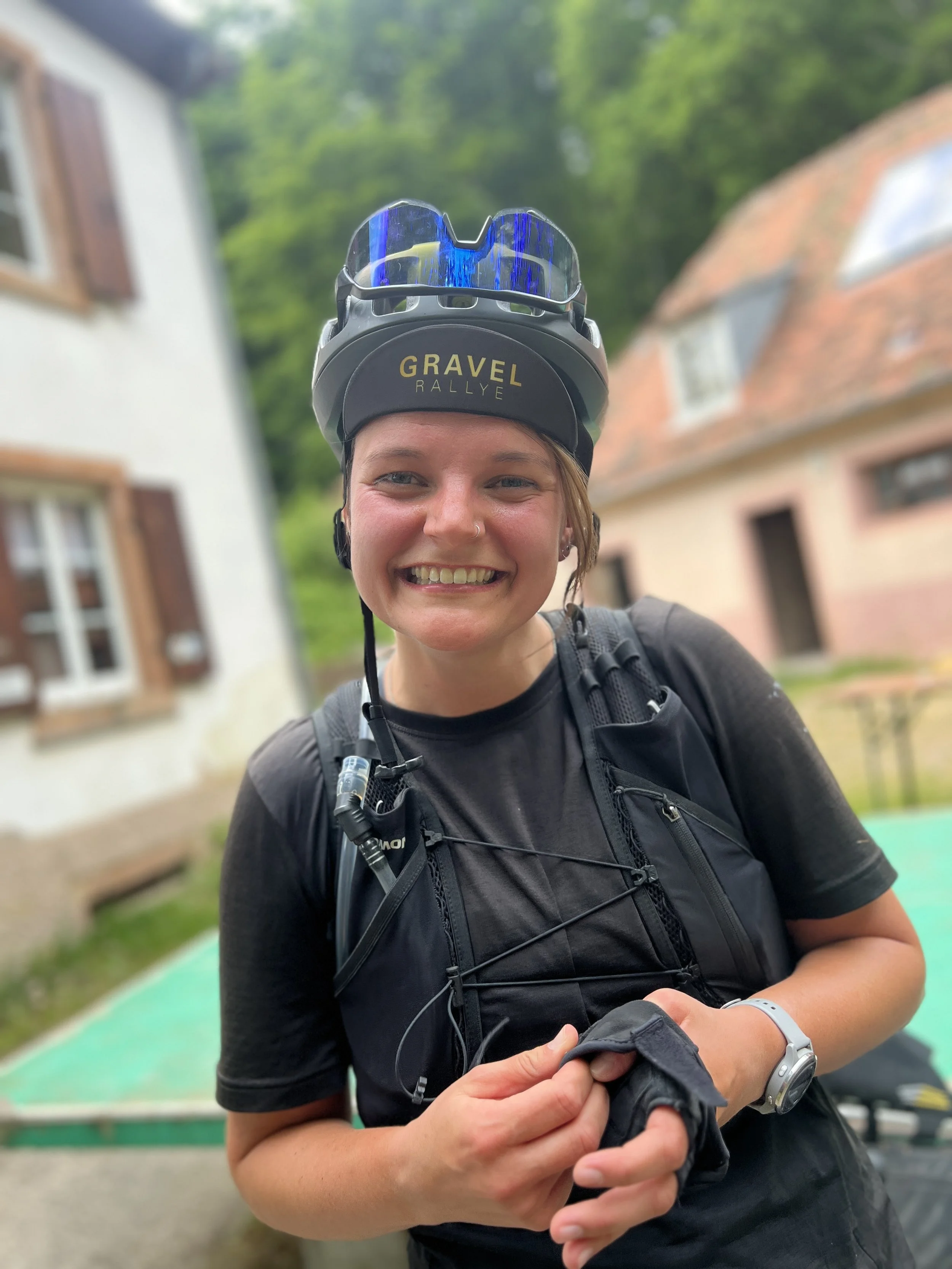 Junge Frau mit Fahrradhelm und Rucksack, lächelnd vor Häusern, draußen in der Natur.