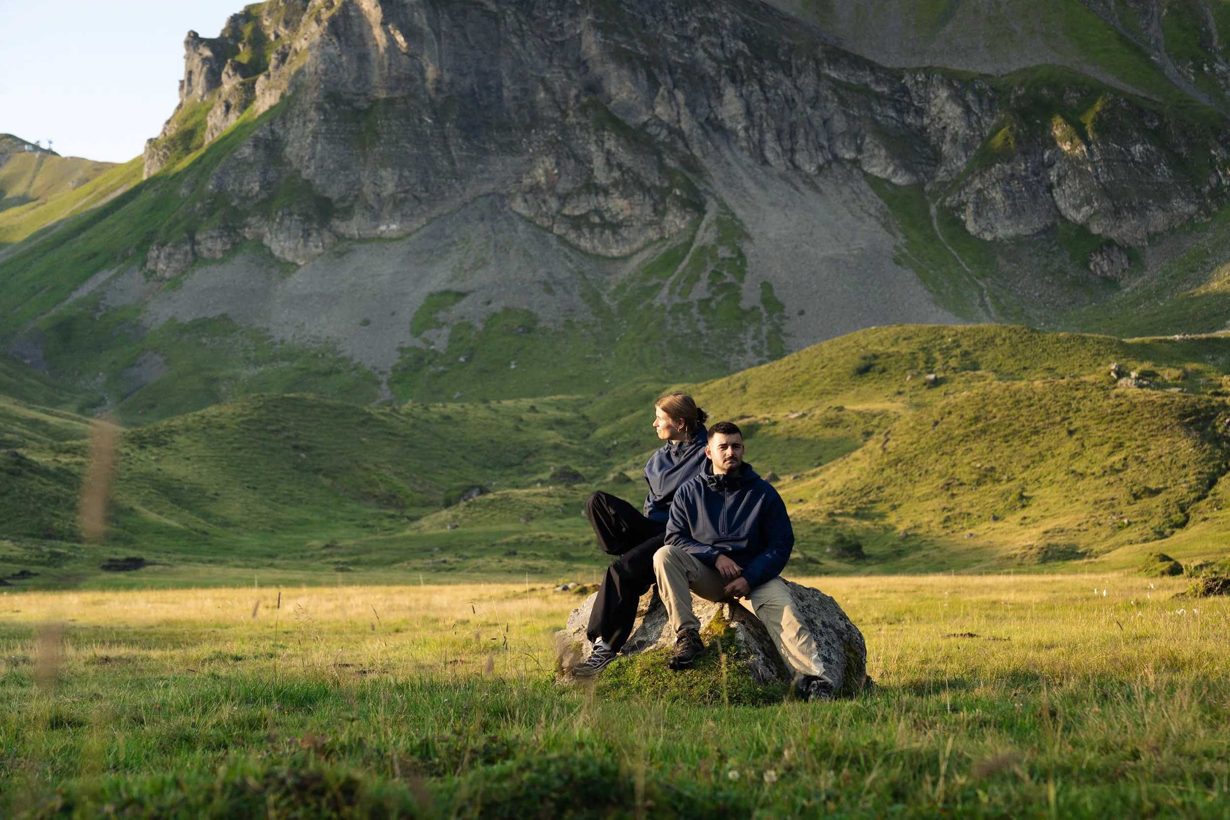 Two people sit on a rock in a green valley with a mountain in the background.nikin. nikin schweiz. flurin giebel. fluringiebel. commercial fotografie. produktefotografie luzern. zentralschweiz.