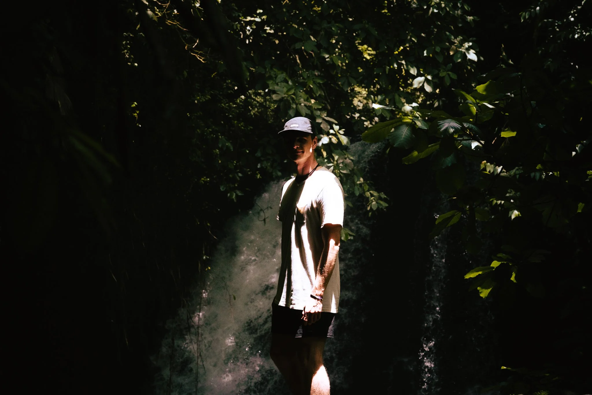 A person wearing a gray cap and a light-colored shirt standing in a dense forest with big green leaves, with sunlight filtering through the canopy, creating a misty and shadowy atmosphere. fineart print. rainforest. flurin giebel. 