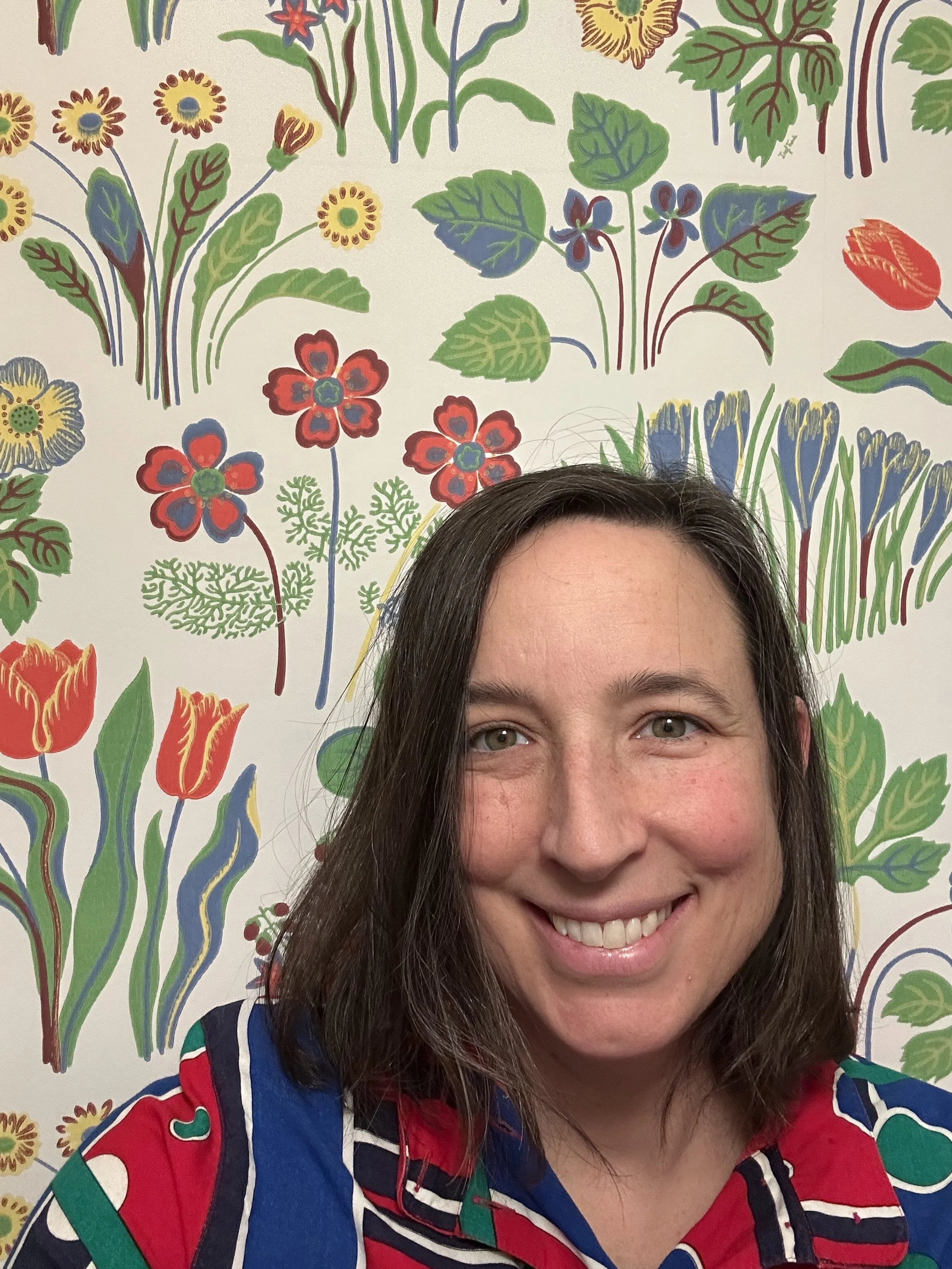 A smiling woman with dark hair in front of a colorful, floral-patterned wall.
