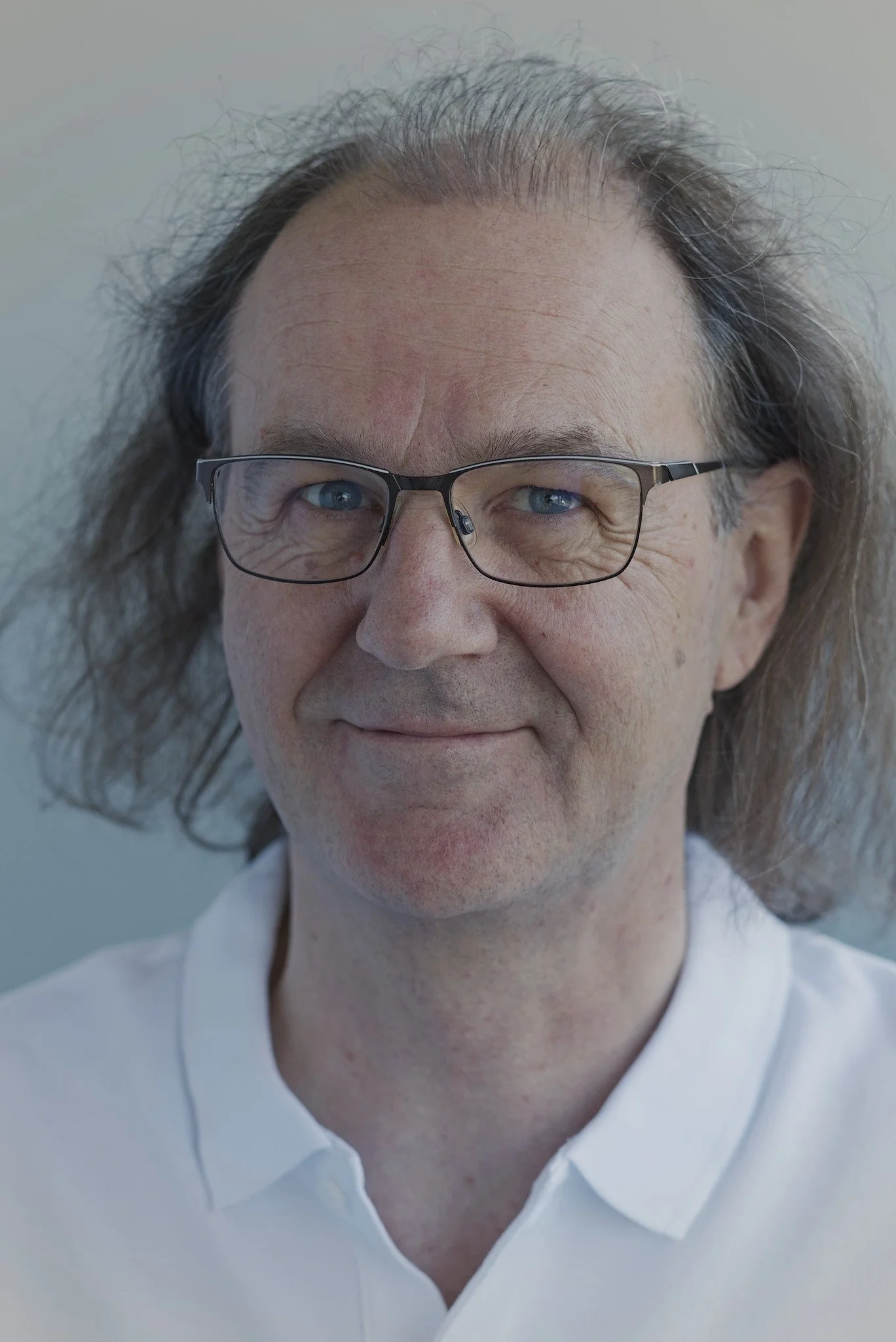 Nahaufnahme eines älteren Mannes mit braunen, lockigen Haaren, Brille und weißem Poloshirt, lächelt leicht. Selbstportrait