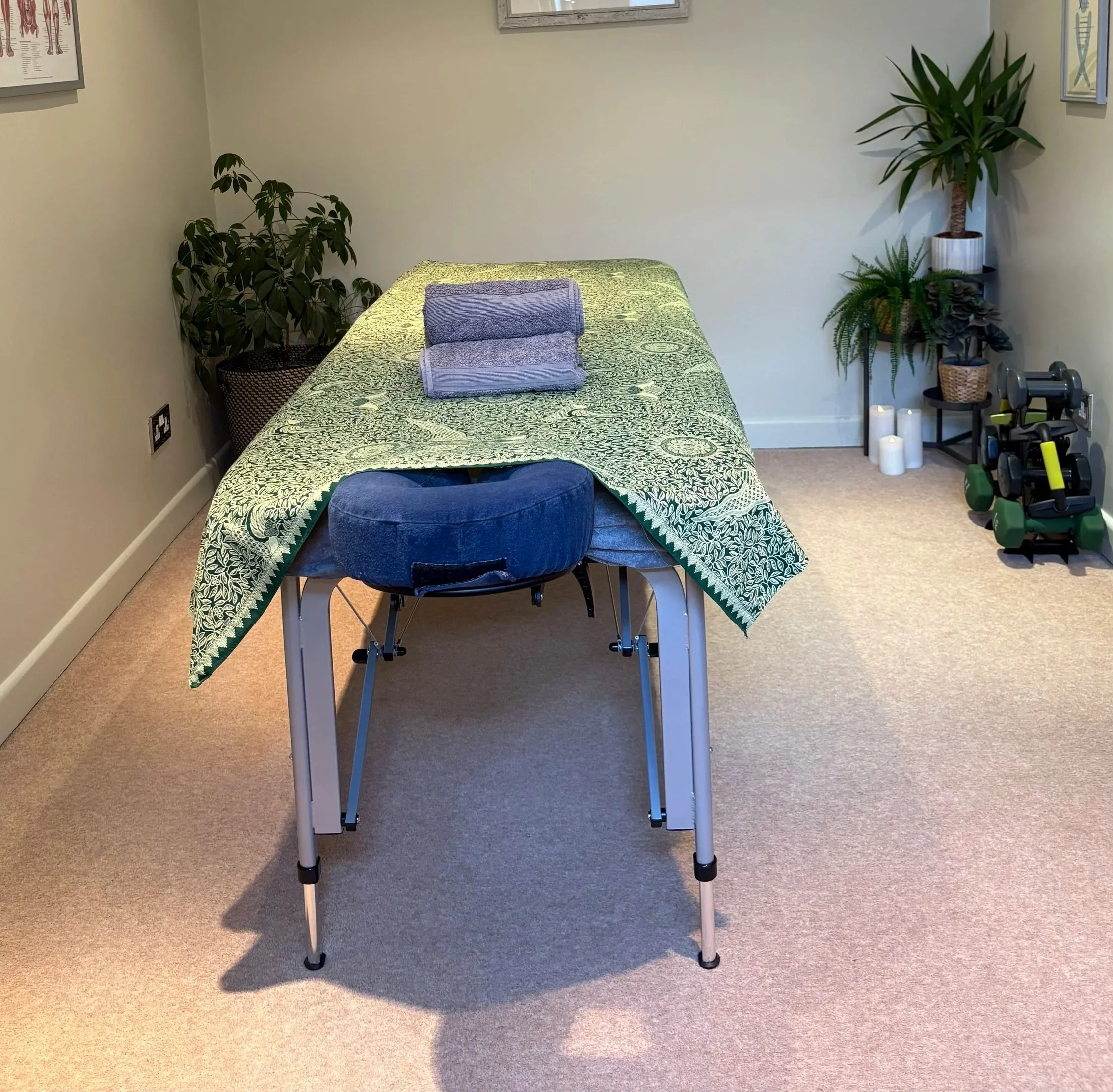 Therapeutic Massage or therapy room with a massage table covered by a green and white patterned cloth, folded towels on top, a reclining stool, potted plants, candles, and some fitness equipment in the corner.