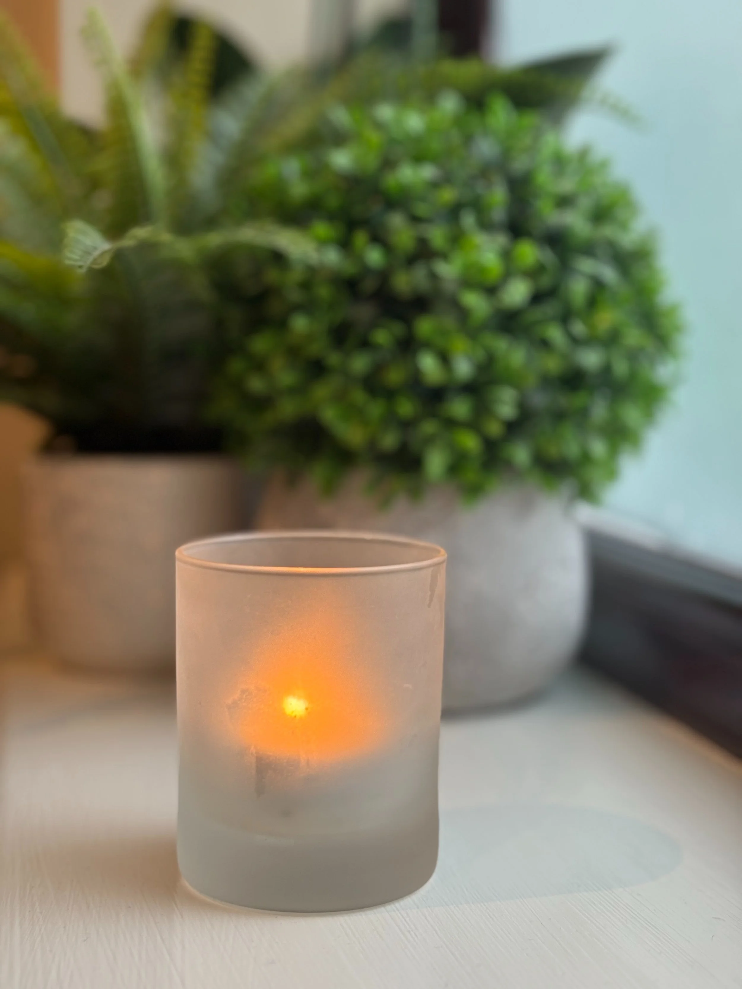 Frosted glass candle holder with a lit candle inside, placed on a white surface near green potted plants in the background.