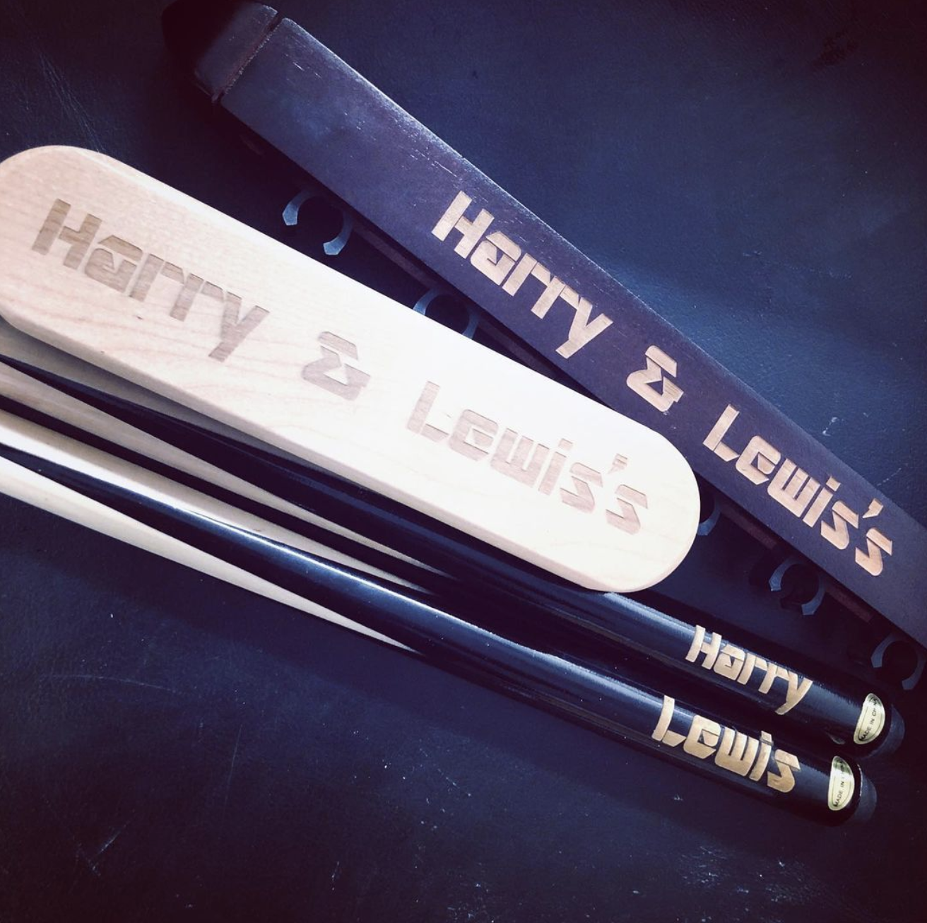 A set of billiard cues and a wooden block engraved with the names 'Henry & Lewis' and 'Harry & Lewis' on a black surface.