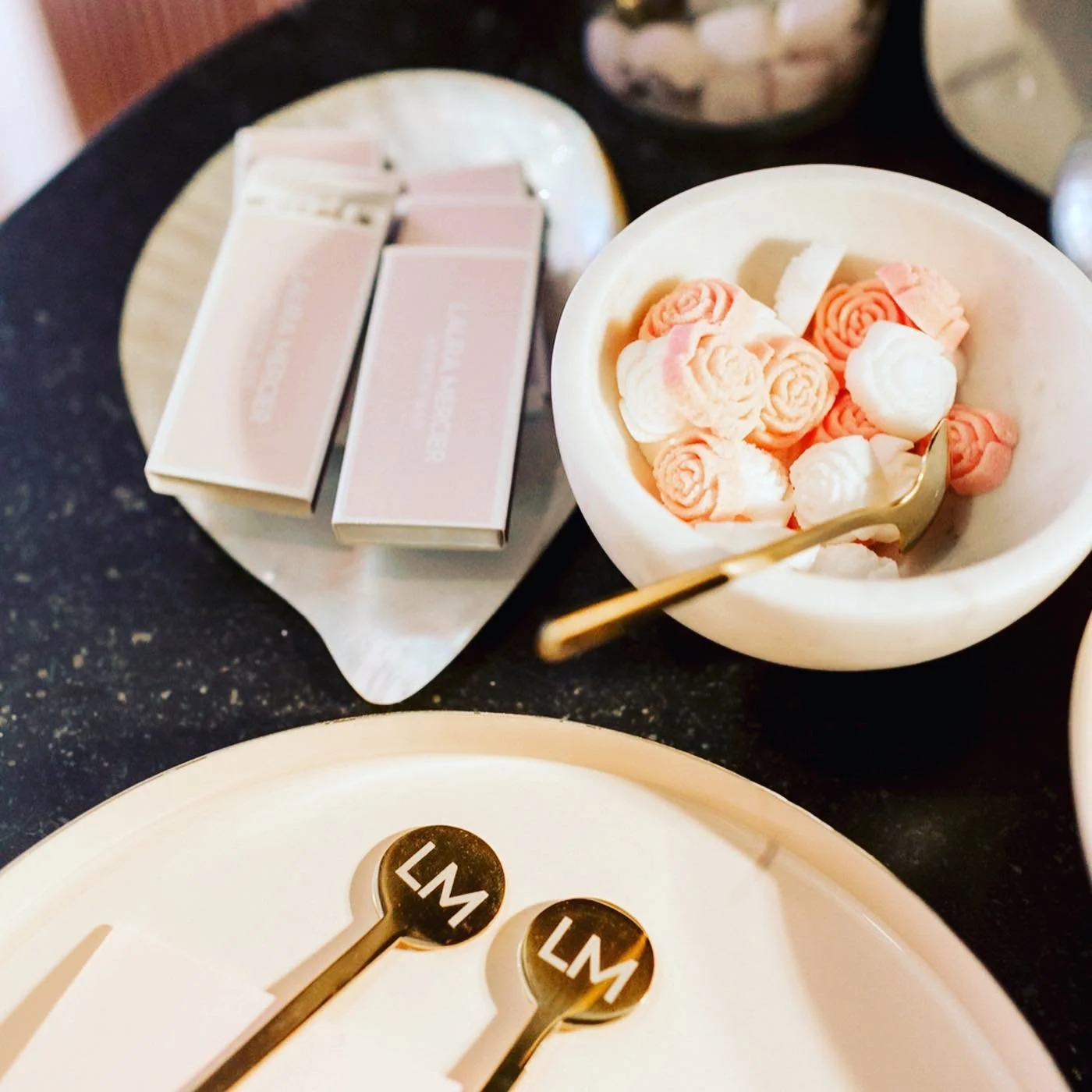 A bowl of soft pink and white rose-shaped marshmallows, a plate with two gold spoons engraved with 'LM', and a tray with pink and white packaged chocolates on a dark countertop.