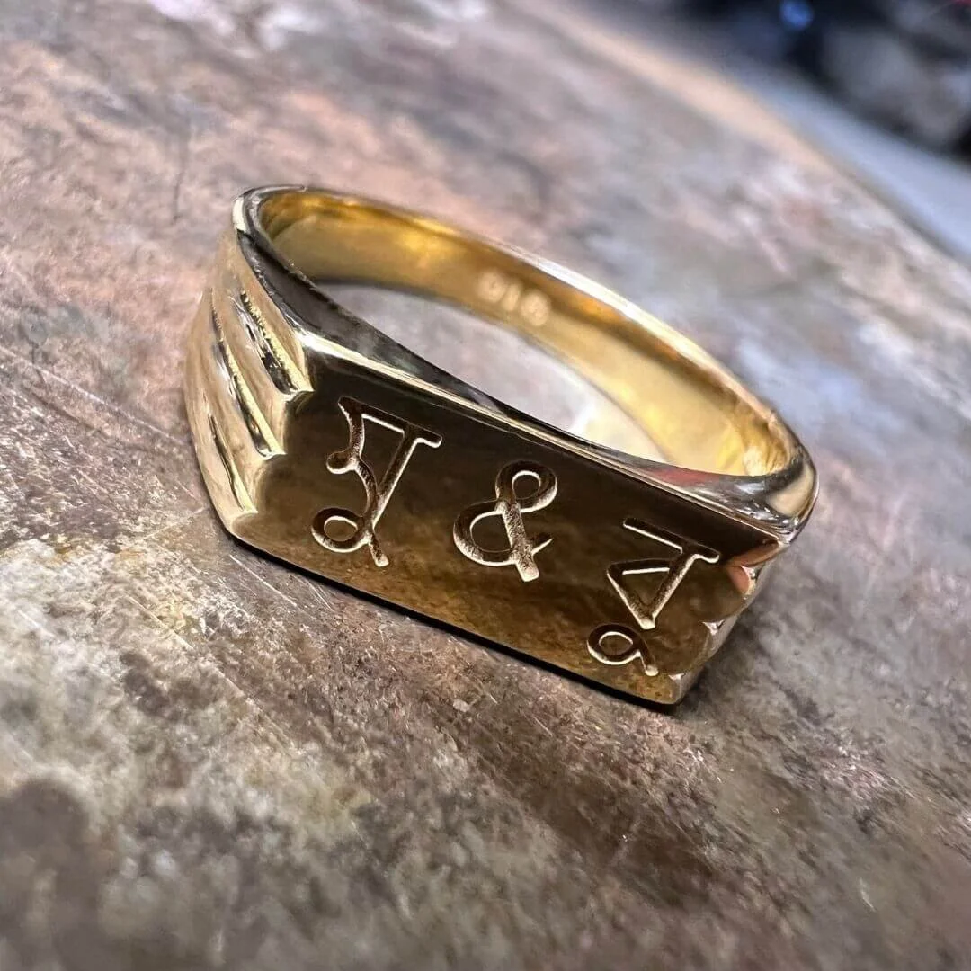 A gold square signet ring engraved with the initials 'J & V' on its face, placed on a jeweller's bench.
