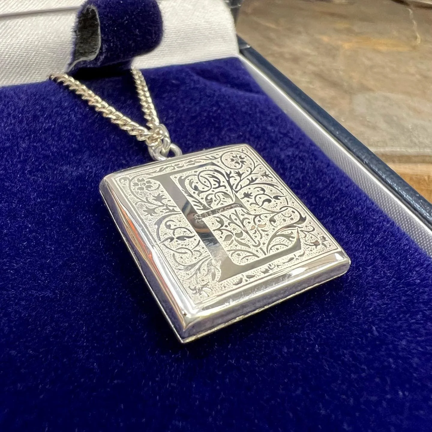 A silver pendant necklace with an intricate engraved design and a large letter 'L' in the center, resting on a blue velvety surface inside a jewelry box.