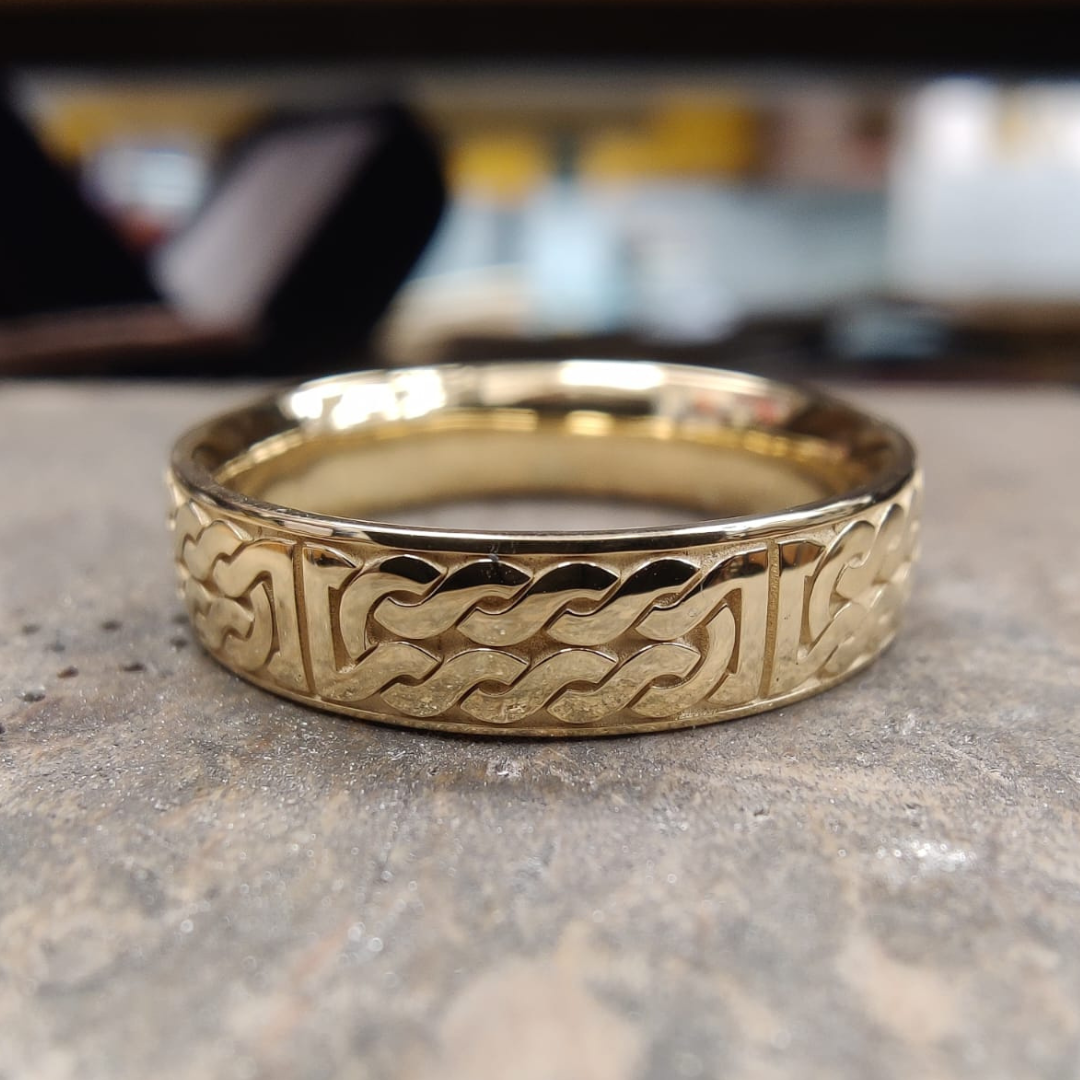 A gold wedding ring engraved with a Celtic knot pattern resting on a wooden jeweller's bench.