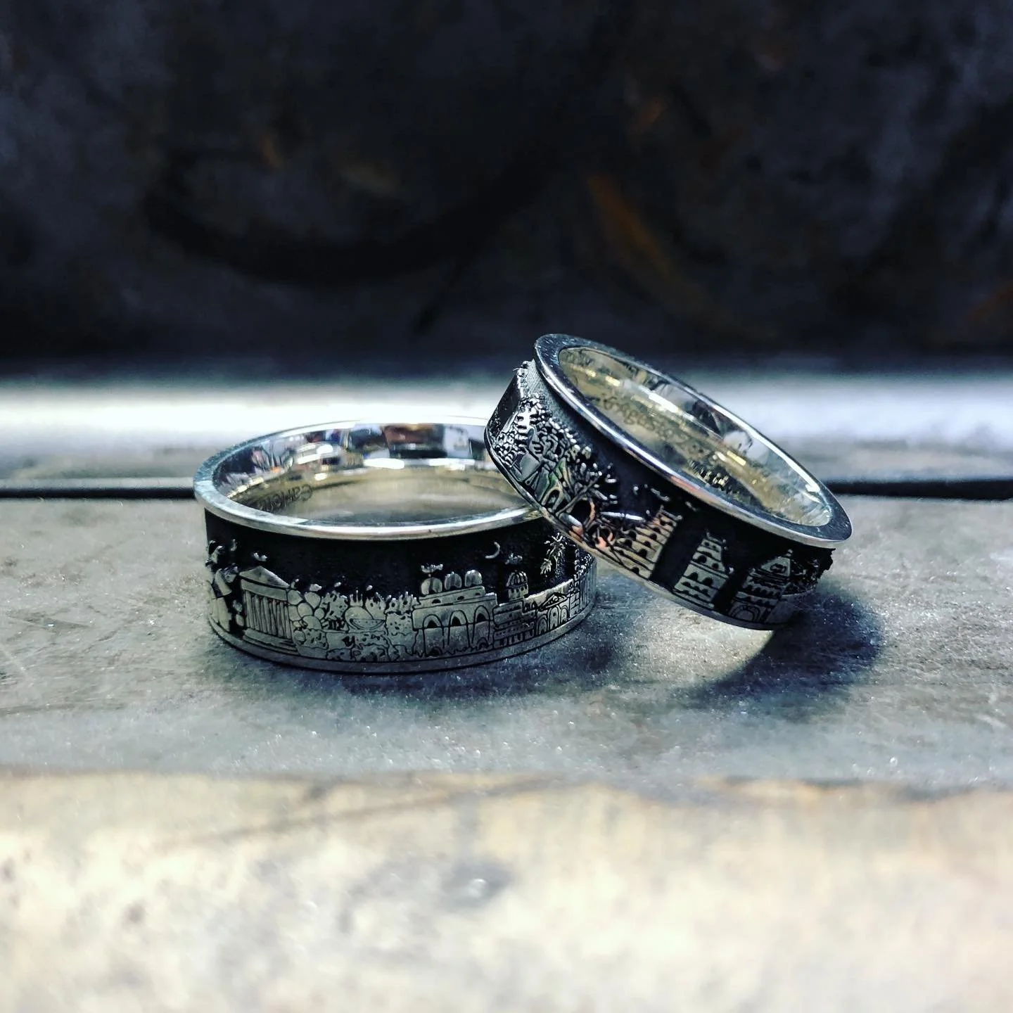 Two silver rings with engraved cityscape and architectural designs resting on a concrete surface, with a dark background.