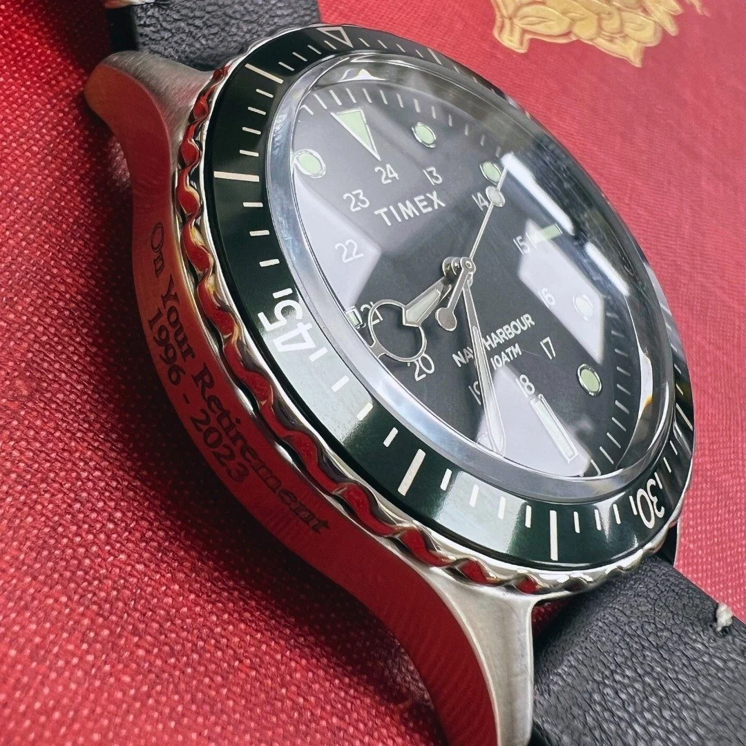 A close-up of an engraved black Timex Navi Harbour watch with a stainless steel case and black leather strap, placed on a red textured surface.