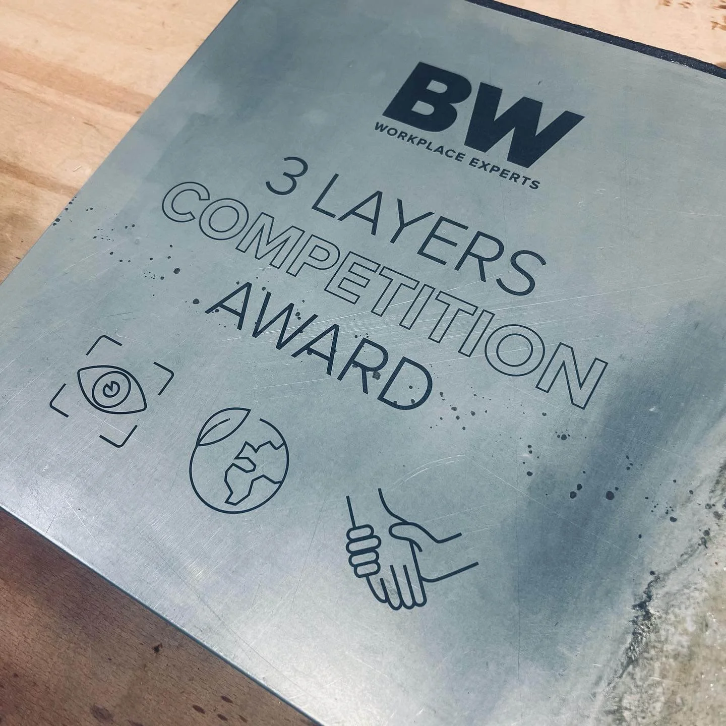 A metallic plaque engraved with Workplace Experts (BW) detailing their 3-layered competition award, featuring icons of an eye, Earth, and a handshake.