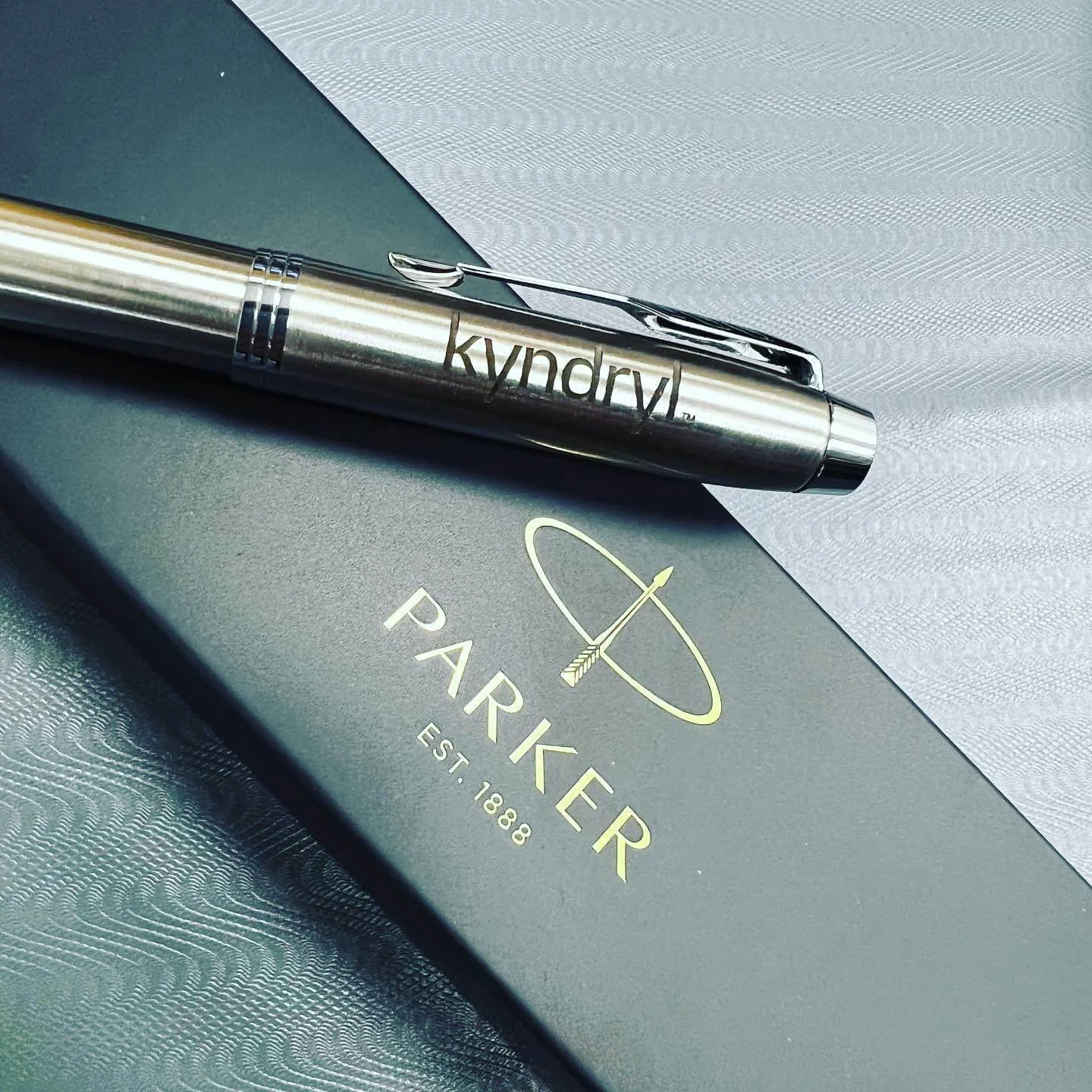 A silver pen with the word 'kyndryl' engraved on it resting on a Parker pen box with the Parker logo and text 'EST. 1888' on it, placed on a textured surface with a paper napkin nearby.