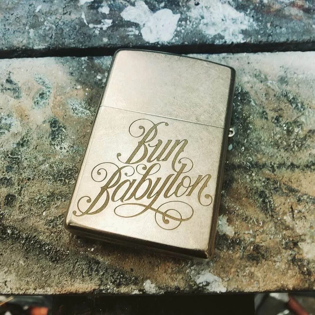 A metallic lighter with the words 'Burn Baby Burn' laser engraved in elegant cursive font on its surface, lying on a rough, textured surface.