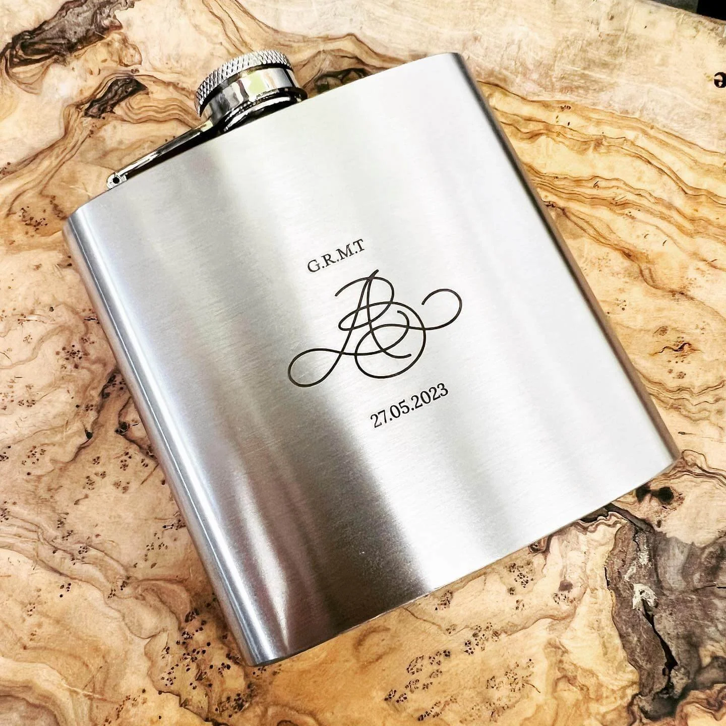 A personalized silver flask with engraved initials and a date, resting on a wooden surface.