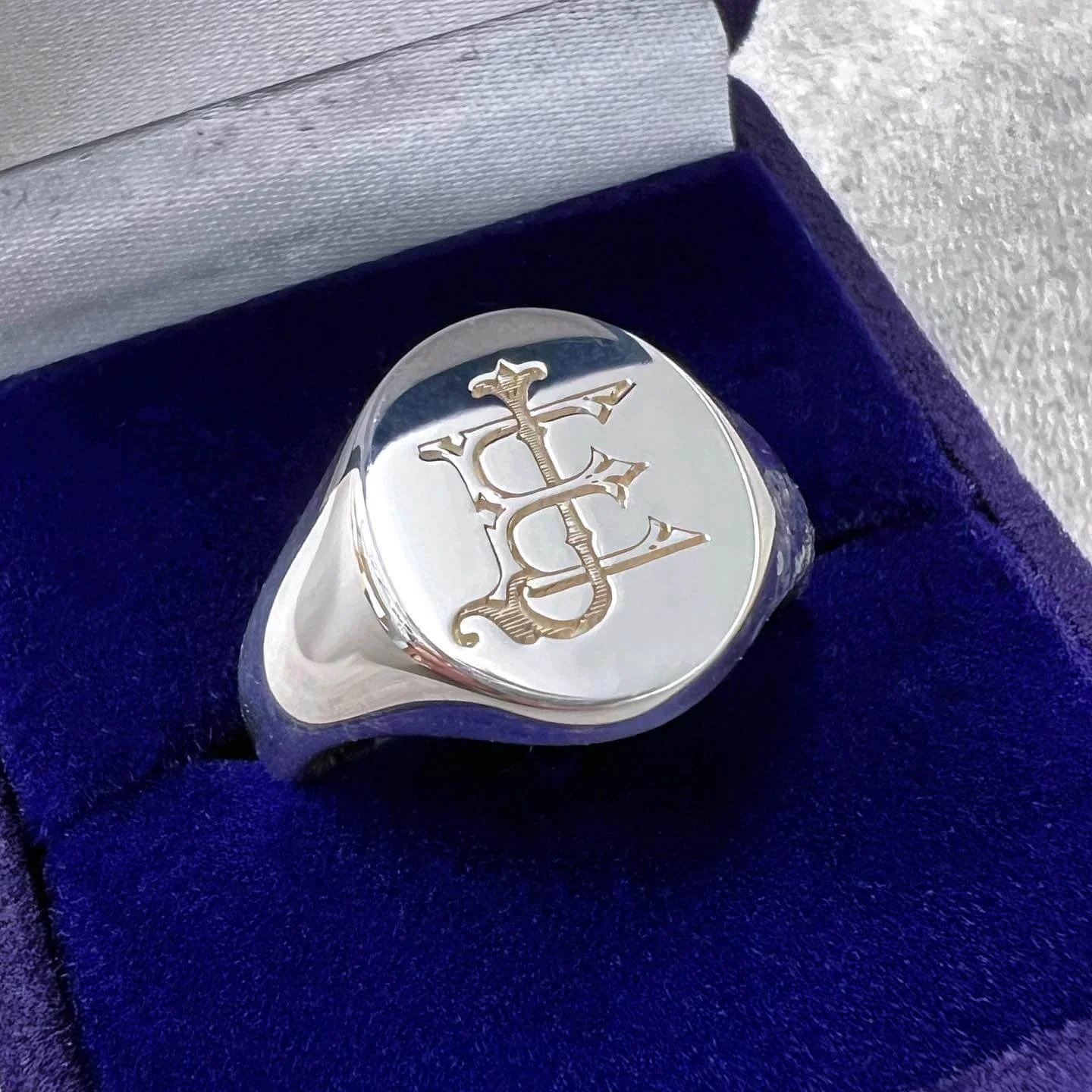 Silver ring with a monogram engraved, resting in a blue jewellery box lined with dark blue fabric.