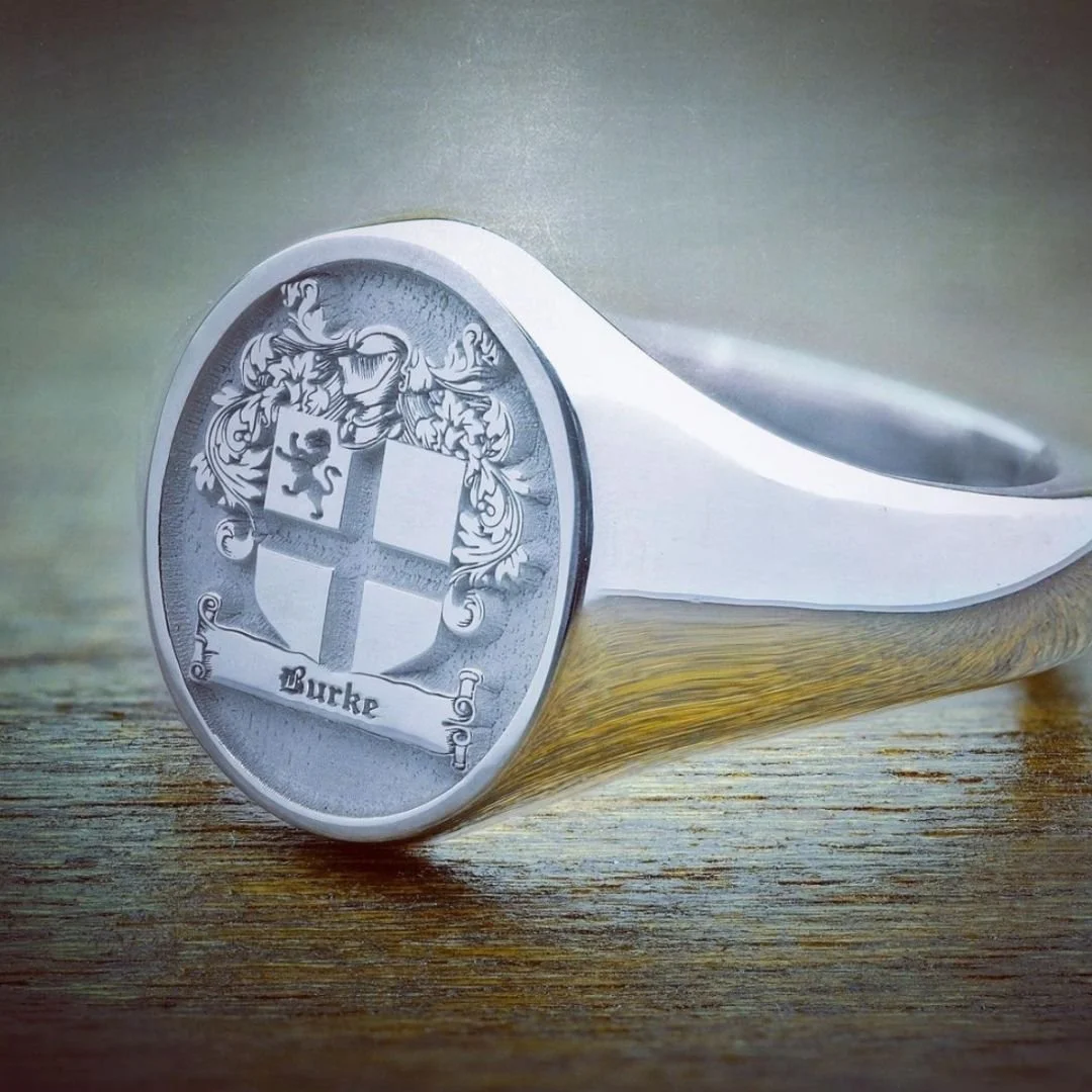 Close-up of a silver signet ring with an engraved crest featuring a lion's head and decorative elements, and the name 'Burke' engraved below.