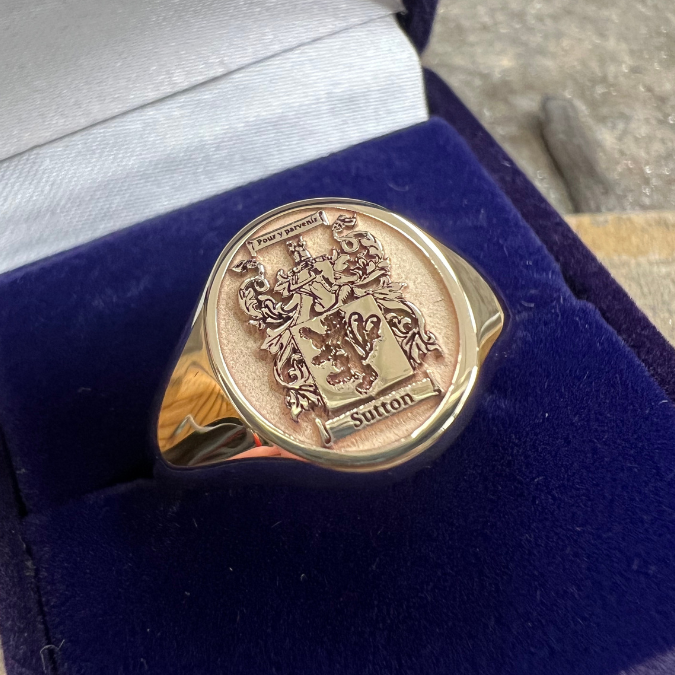 A gold signet ring with a detailed engraved crest and the name 'Sutton' engraved, resting in a blue velvet box.