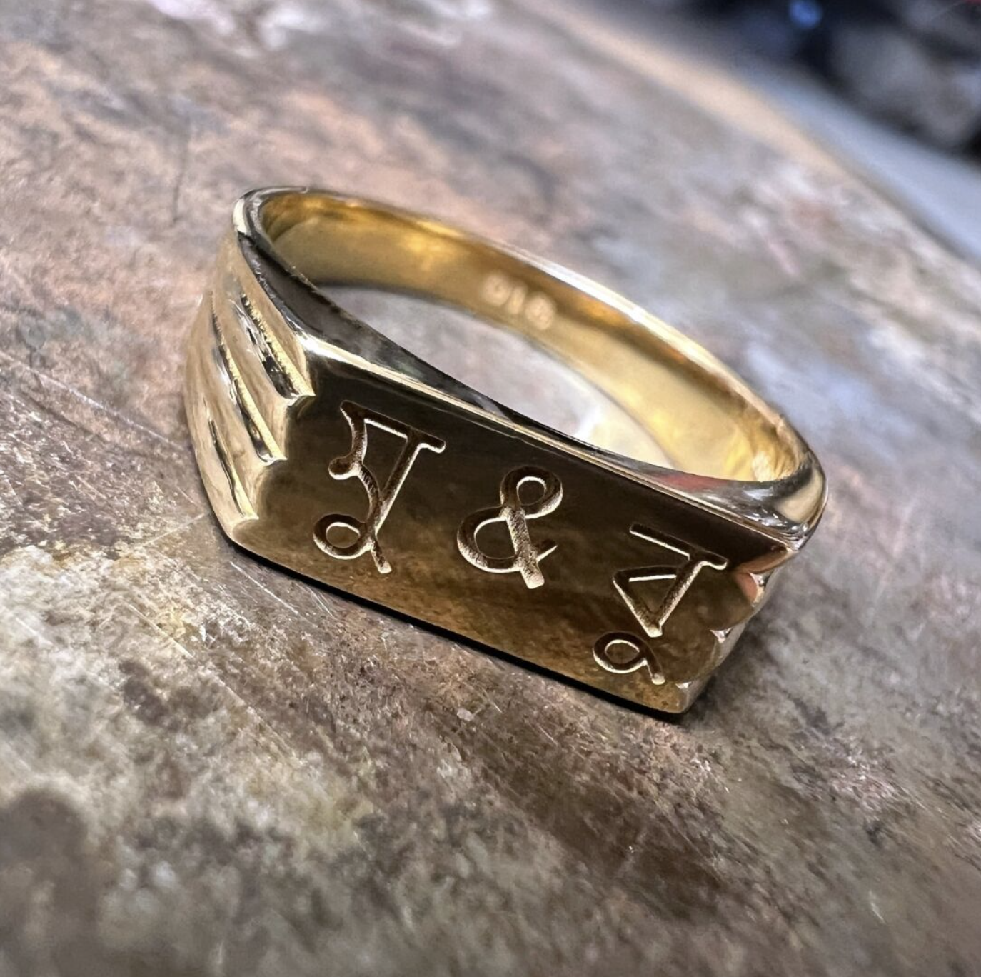 A Gold square signet ring engraved with the initials 'V & J' resting on a jeweller's bench.