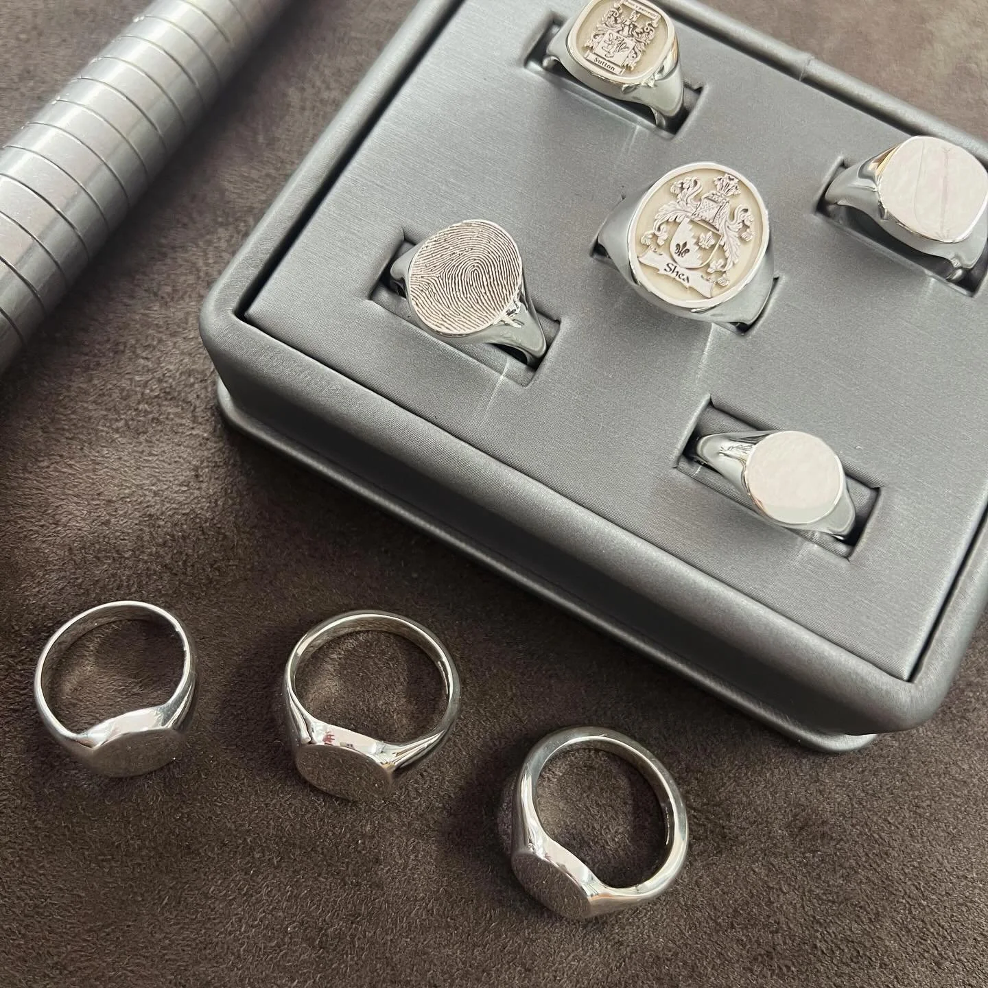 Set of four silver signet rings with different designs on a dark surface, with a grey box holding some rings, including one with a fingerprint design and another with a crest, and a silver cylindrical object to the side.