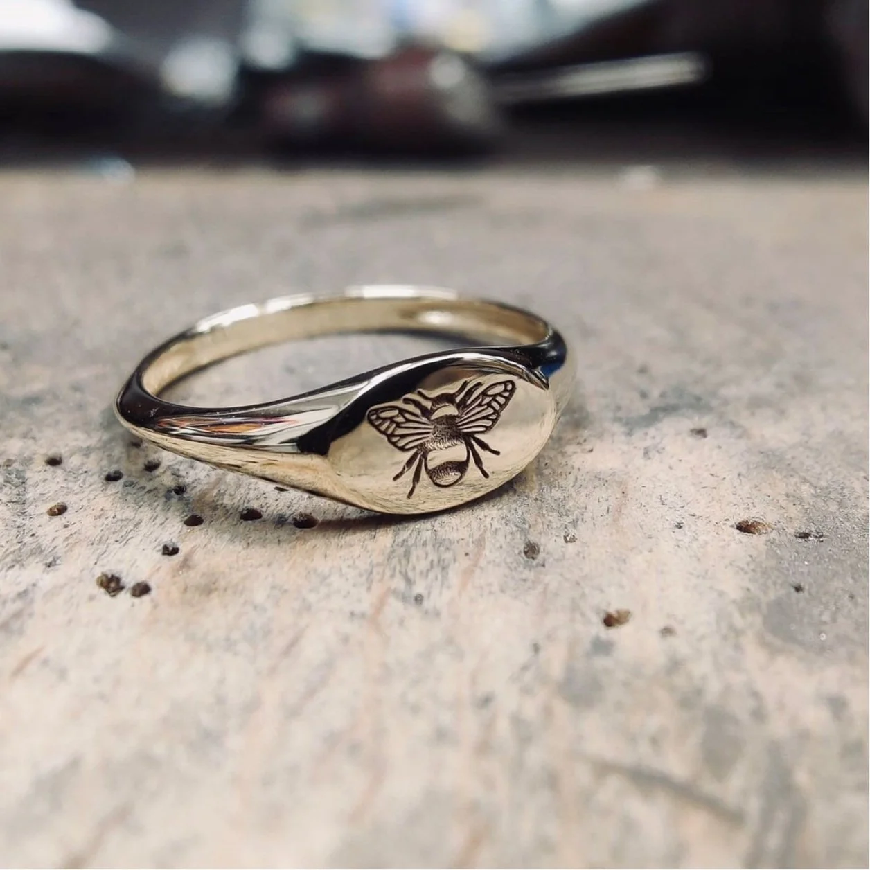 A silver ring with a engraved bee on its surface, resting on a rough gray surface.