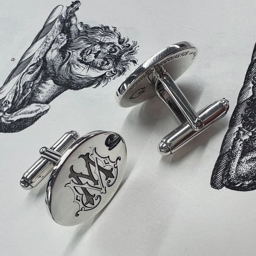 A pair of silver cufflinks with a black and white crest design that has been laser-engraved is placed on paper with illustrations of a lion and a bear.