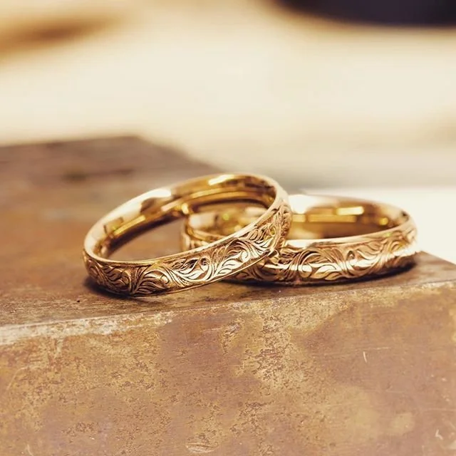 Two gold wedding rings with engraved floral patterns resting on a wooden surface.