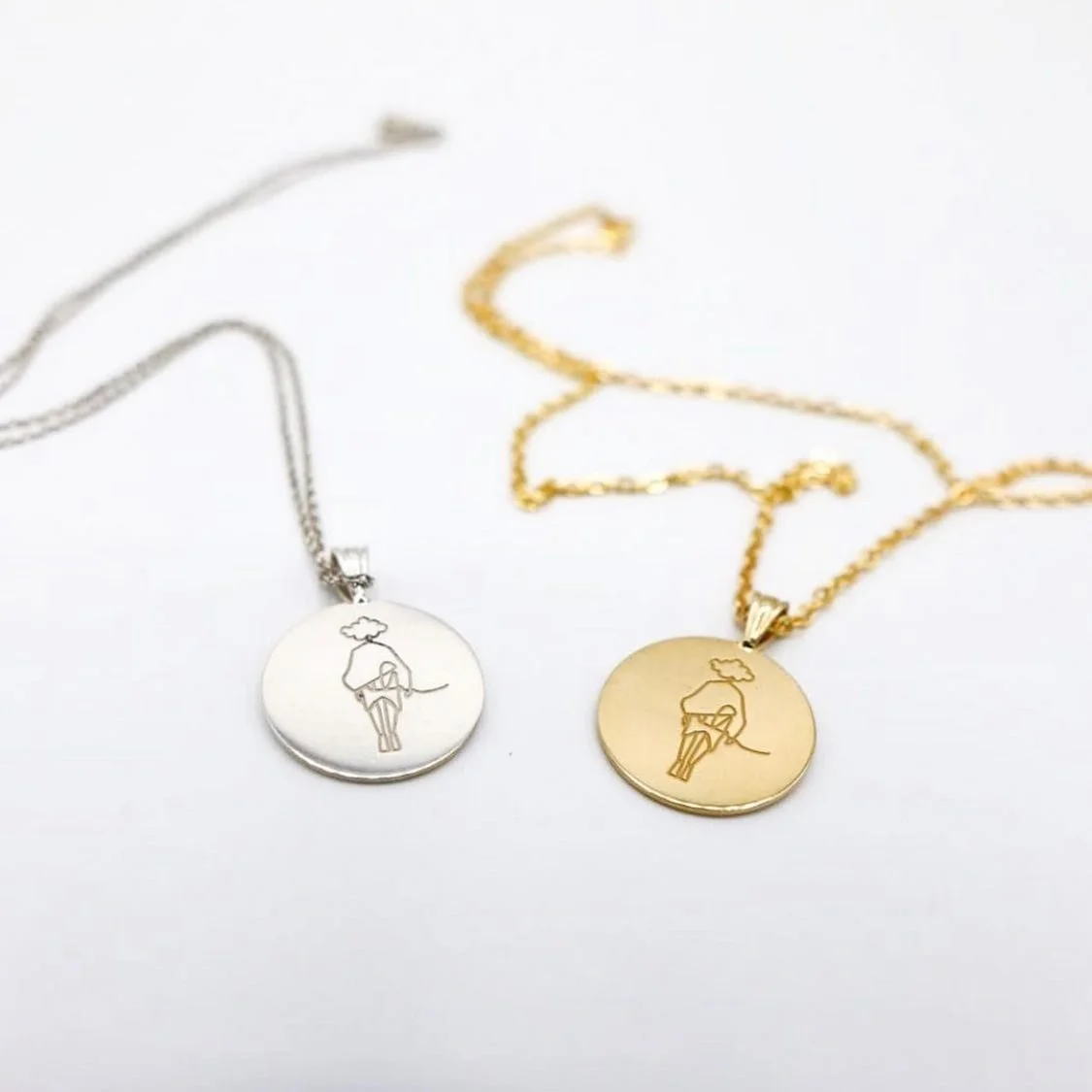 Two round pendant necklaces, one silver and one gold, each engraved with a stylised woman, lying on a white surface.