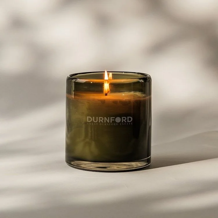 A lit candle in a dark glass container with the words 'DURNORD' and 'GREAT DURNORD ESTATE' engraved on it, placed on a white surface with a blurred background.
