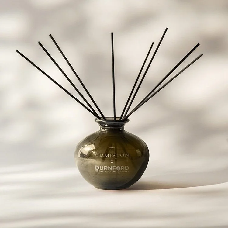 A brown glass vase with reed diffuser sticks inside, placed against a blurred light background. The vase features branding that has been engraved, reading 'Edmiston x Durnford, Great Durnford Estate'.