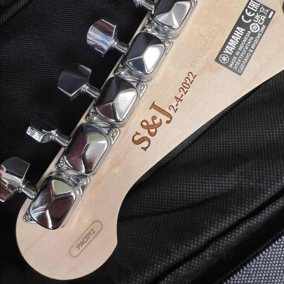 Close-up of the headstock of a Yamaha guitar with tuning pegs and a natural wood finish, engraved with the initials 'SJ&J' and the date '2-4-2022. '