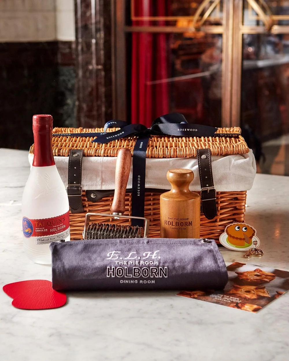 A wicker picnic basket with black straps on a white marble table, surrounded by a bottle of gin, a wooden pepper grinder, a gray pouch with white text, two red paper hearts, and a small cartoon chicken sticker, with a glass of dark drink in the backg