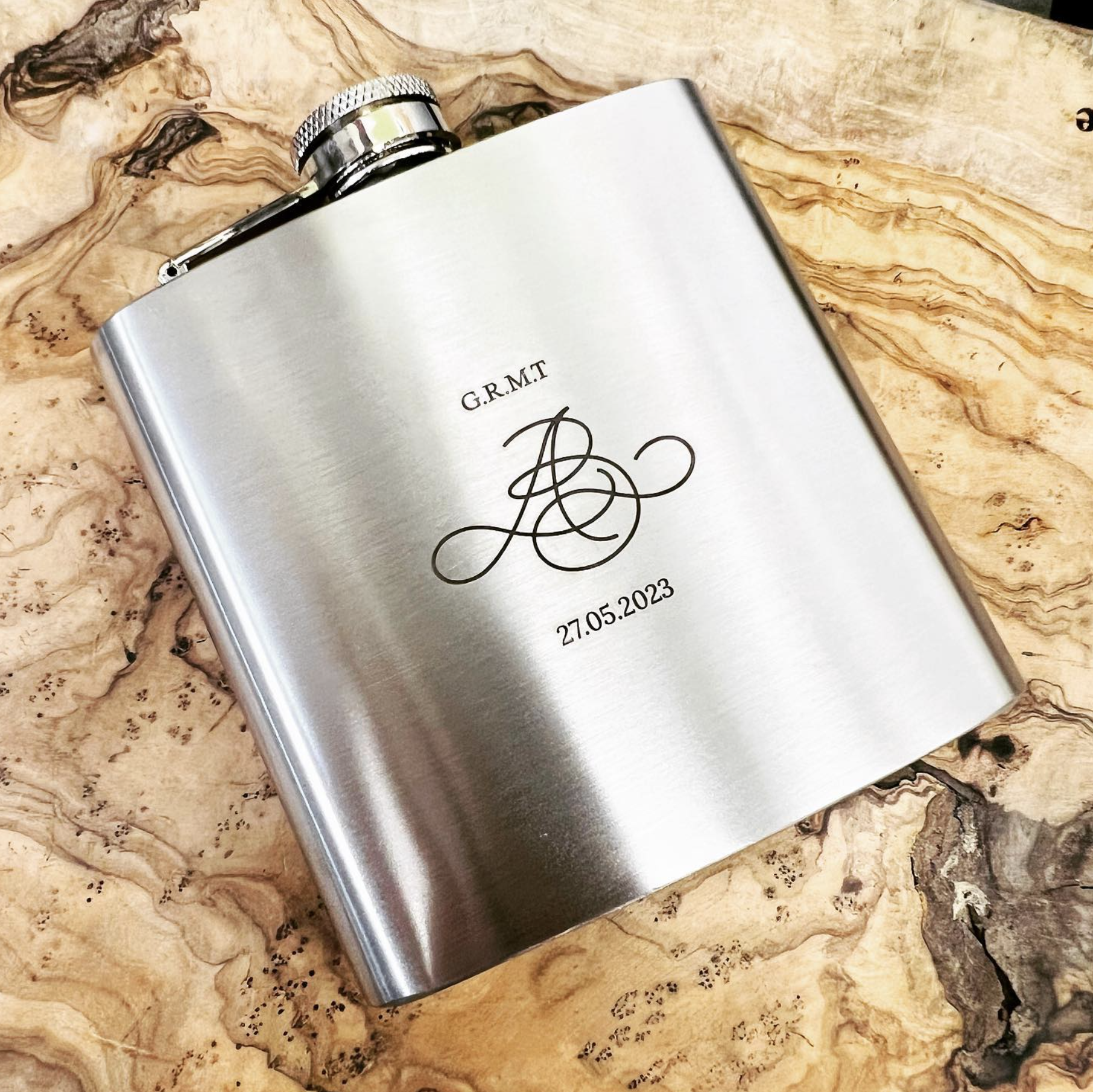 A silver flask with engraved initials, date, and decorative monogram placed on a wooden surface.