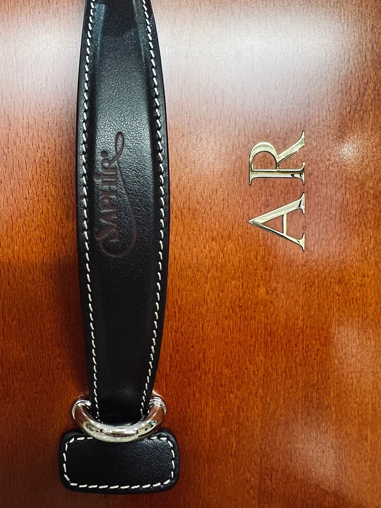 A black leather strap with white stitching, labeled 'CONAIR', resting on a wooden surface next to a shiny brass 'A' and 'R' letter that has been laser cut out of brass.