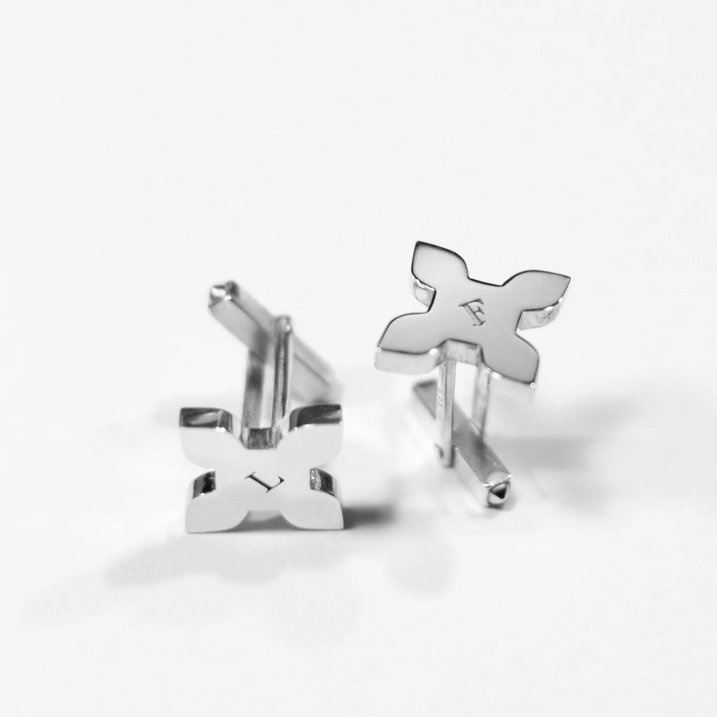 Pair of silver cufflinks with cross-shaped designs and engraved letter 'L' on each, resting on a white surface.