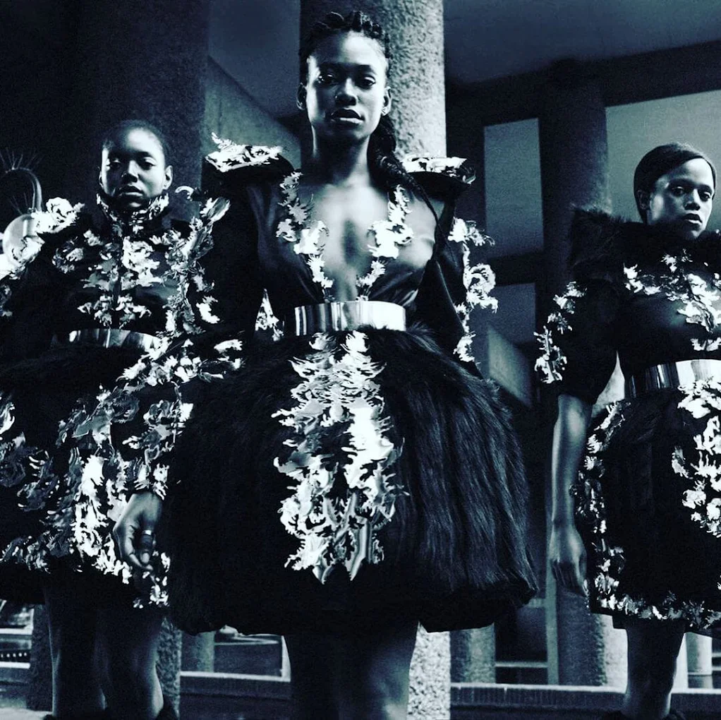 Three women wearing elaborate black dresses with white floral patterns that have been laser cut, standing indoors against a dark background.