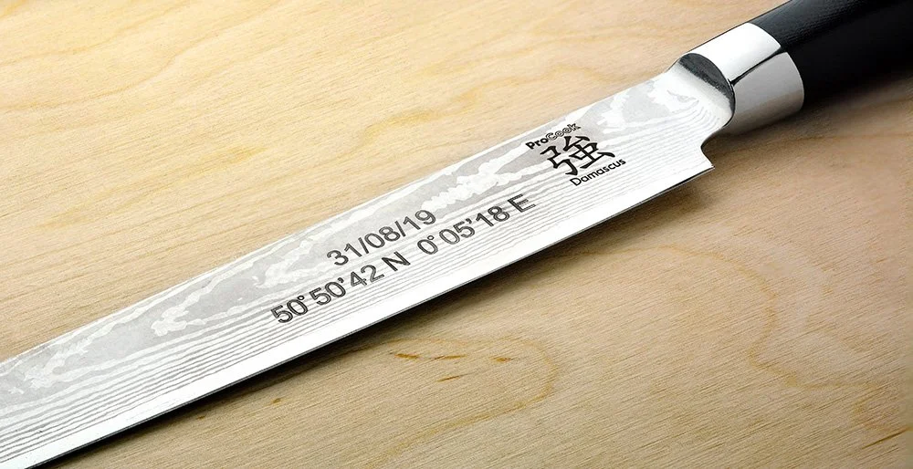 A close-up of a kitchen knife with a black handle, a metal bolster, and a Damascus steel blade, resting on a wooden surface. The blade has been engraved with coordinates, a date, and several symbols.
