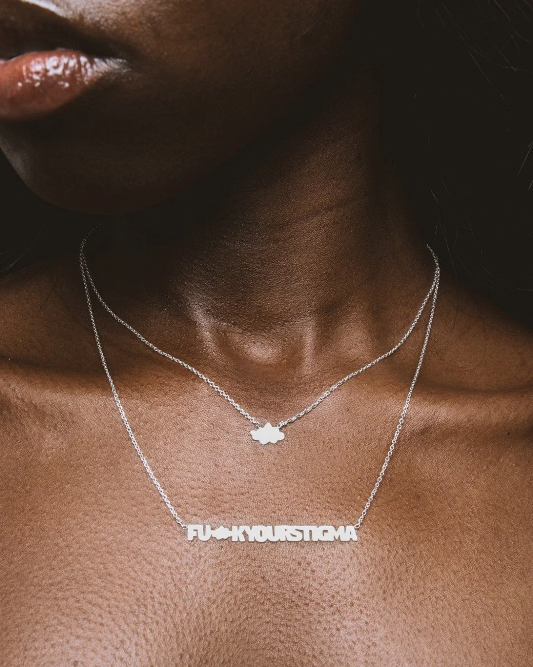 Close-up of a person's neck and chest with two-layered silver necklaces, one with a cloud pendant and the other with text reading 'FU-KYOUSTIGMA', both of which had been laser cut from silver sheet.