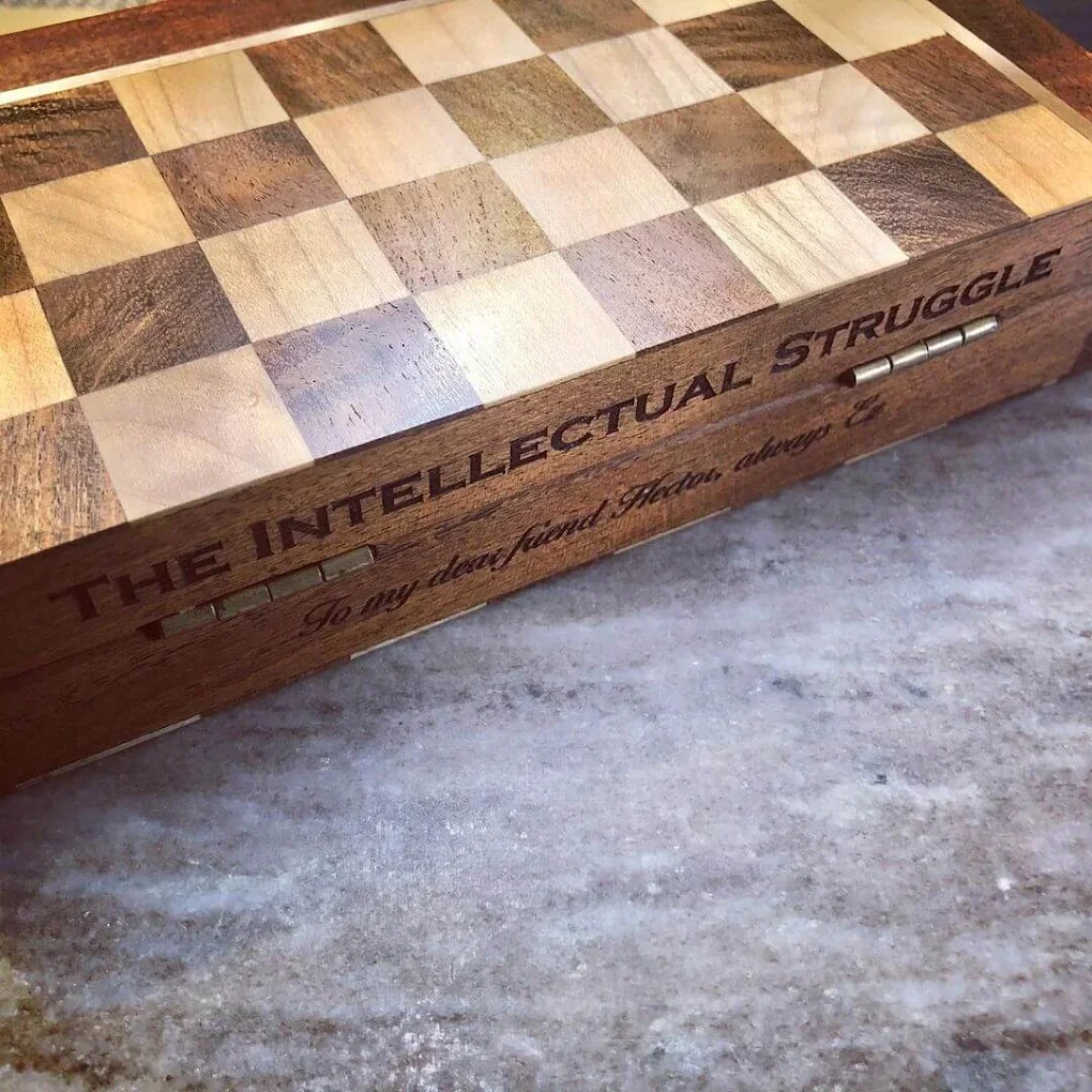 A wooden chessboard with the phrase "The Intellectual Struggle" engraved on the side, and a hinge for opening or closing.
