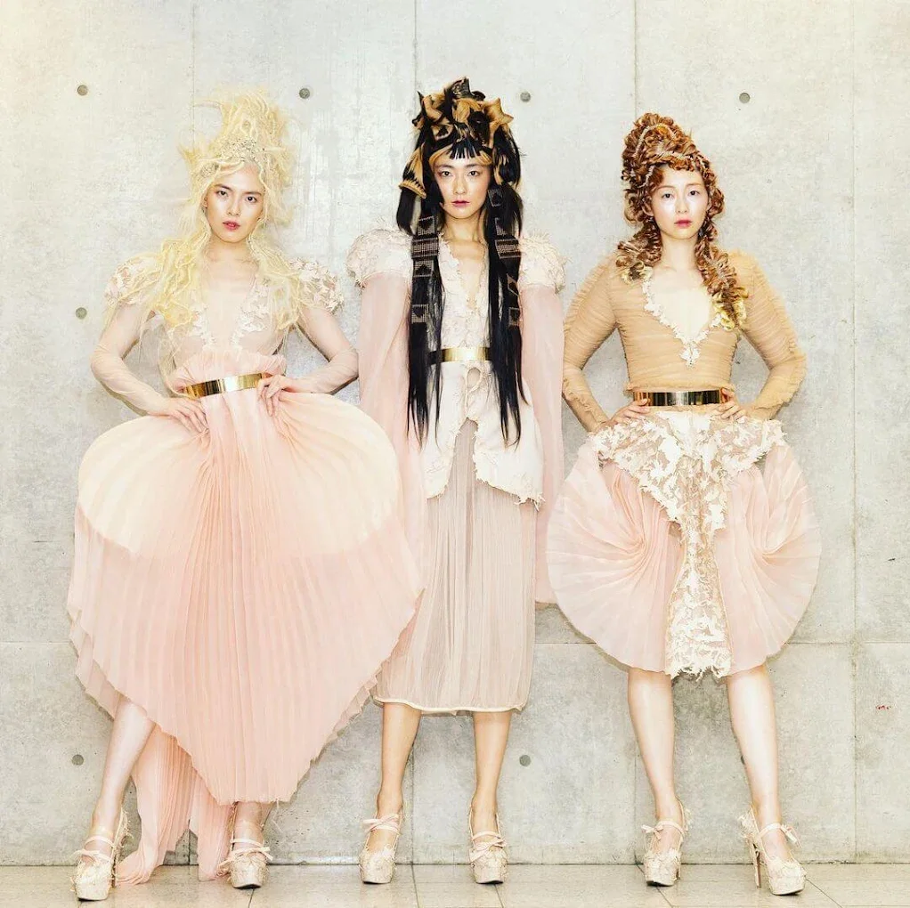Three women standing against a concrete wall, with intricate hairstyles, dressed in elaborate, pastel-colored clothing that has been laser cut and hand stitched together.