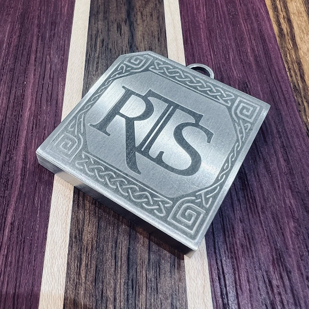 A metallic pendant with the initials 'RKS' engraved in the center, decorated with a Celtic knot border, resting on a multicolored wooden surface.