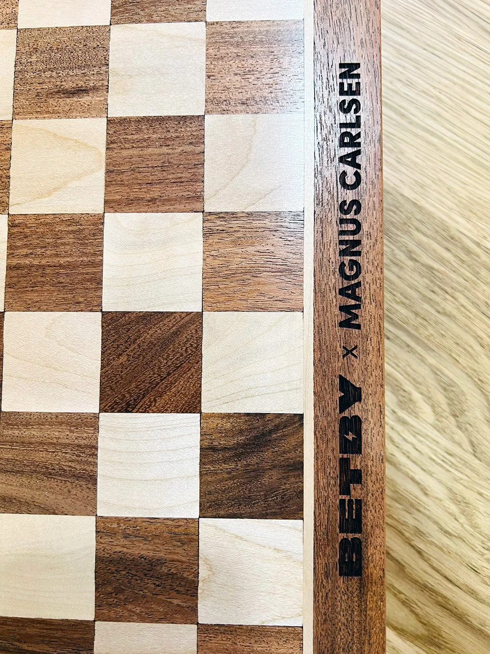 Close-up of a wooden chessboard with alternating light and dark squares, with a wooden border engraved with the text "BETA by X MAGNUS CARESIEN."
